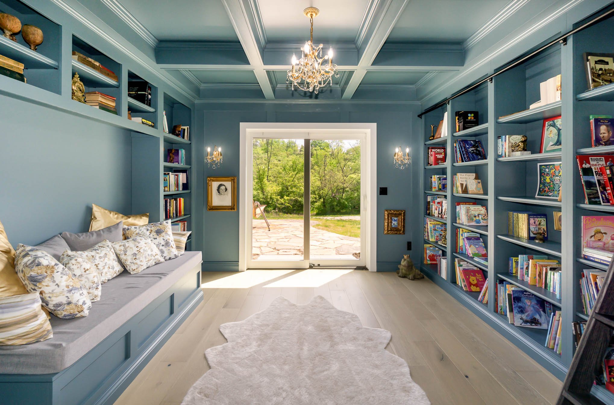 Custom reading room with blue built-in bookshelves filled with colorful books and decorative objects. A window shows greenery outside, and there are chandeliers hanging from the ceiling, with a small lamp on the left side.