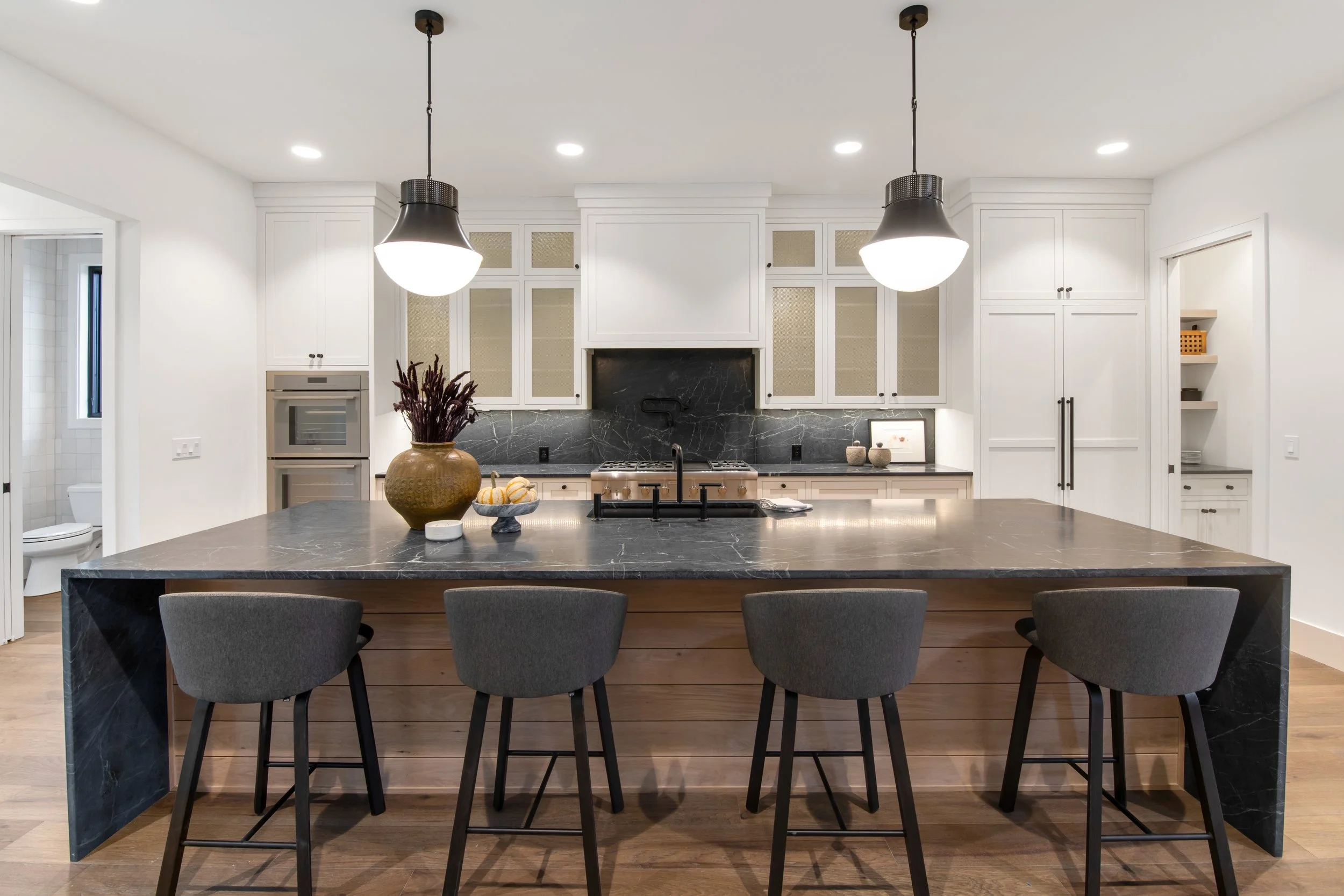 Custom kitchen with white cabinets, black marble backsplash, gray dining chairs, black pendant lights, and a large island with dark stone countertop.