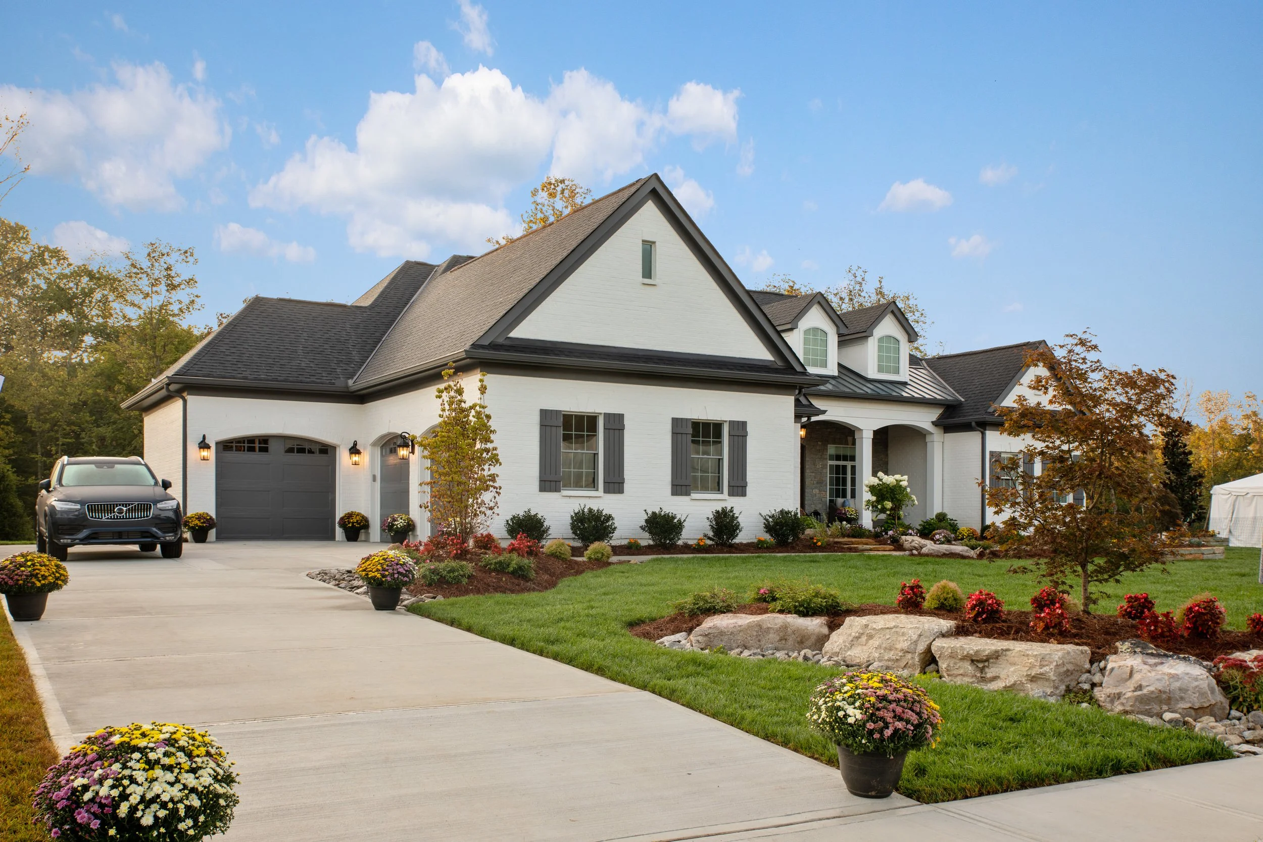 White two-story suburban house with black shutters, gray roof, attached garage, and landscaped front yard with flowers and trees.