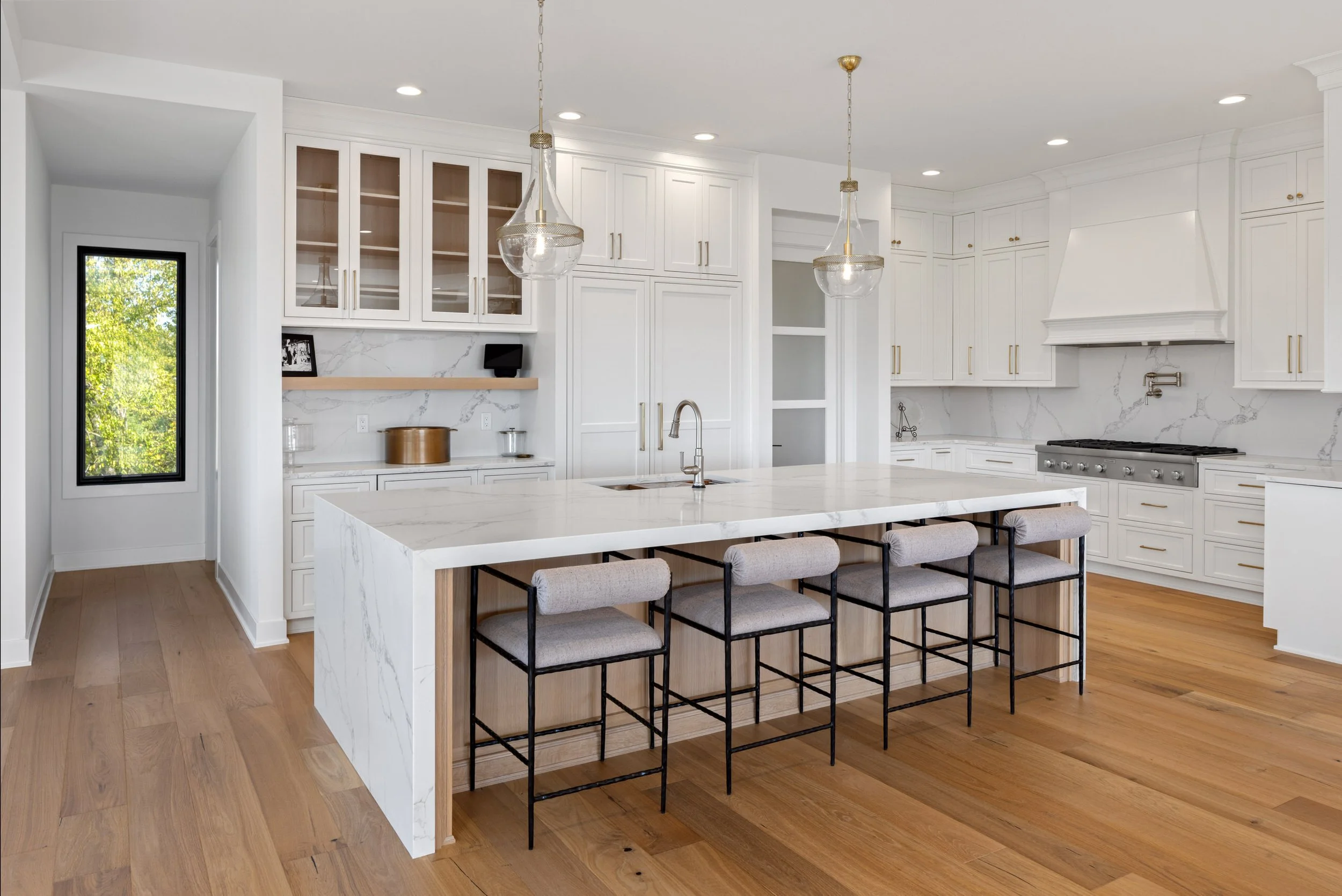 Elegant white kitchen with a large island, bar stools, upper cabinets with glass doors, and hardwood flooring. Two pendant lights hang over the island. There is a window on the left side of the kitchen.