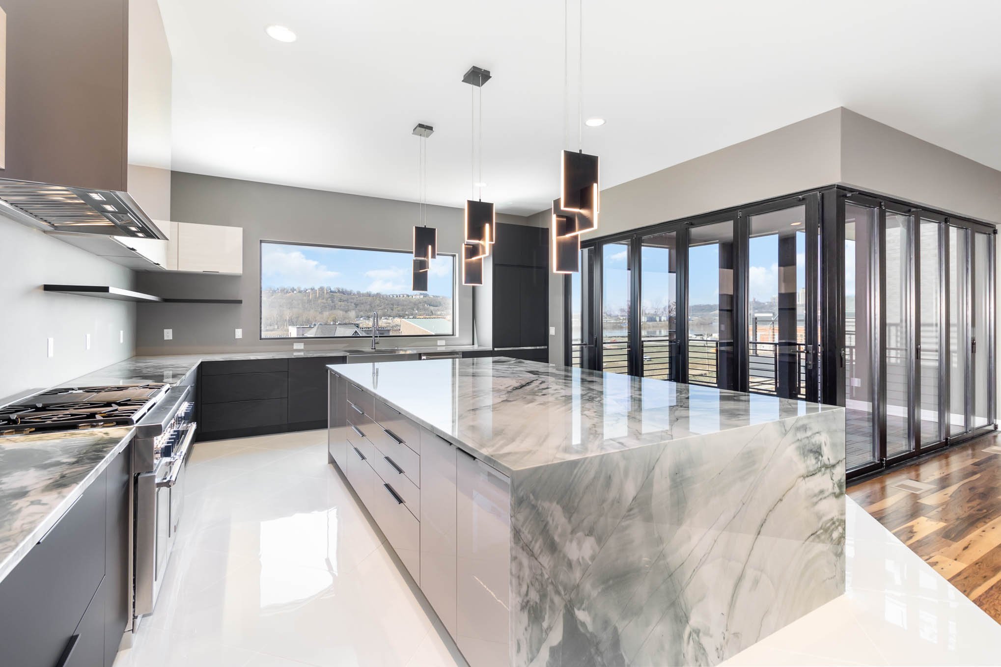 Modern kitchen with marble island, gray cabinets, stainless steel stove, large window, and black pendant lighting fixtures.