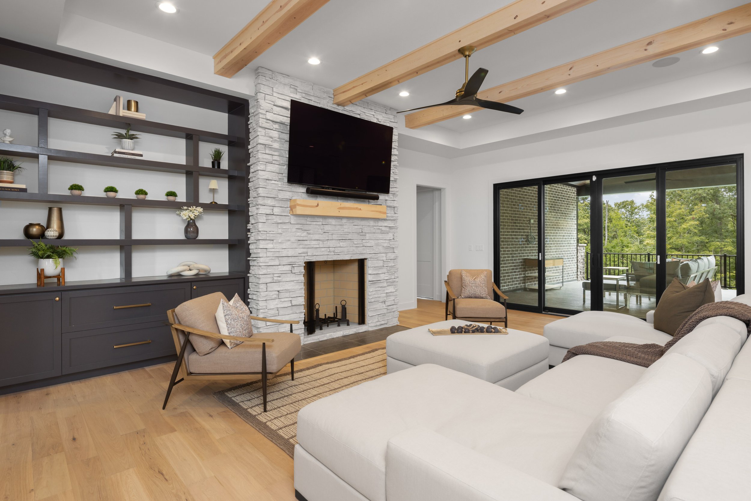 Living room with white sectional sofa, wooden chairs, a gray built-in shelf with plants and decor, a white stone fireplace, a large flat-screen TV, wood beams on the ceiling, and sliding glass doors leading to a balcony with trees outside.