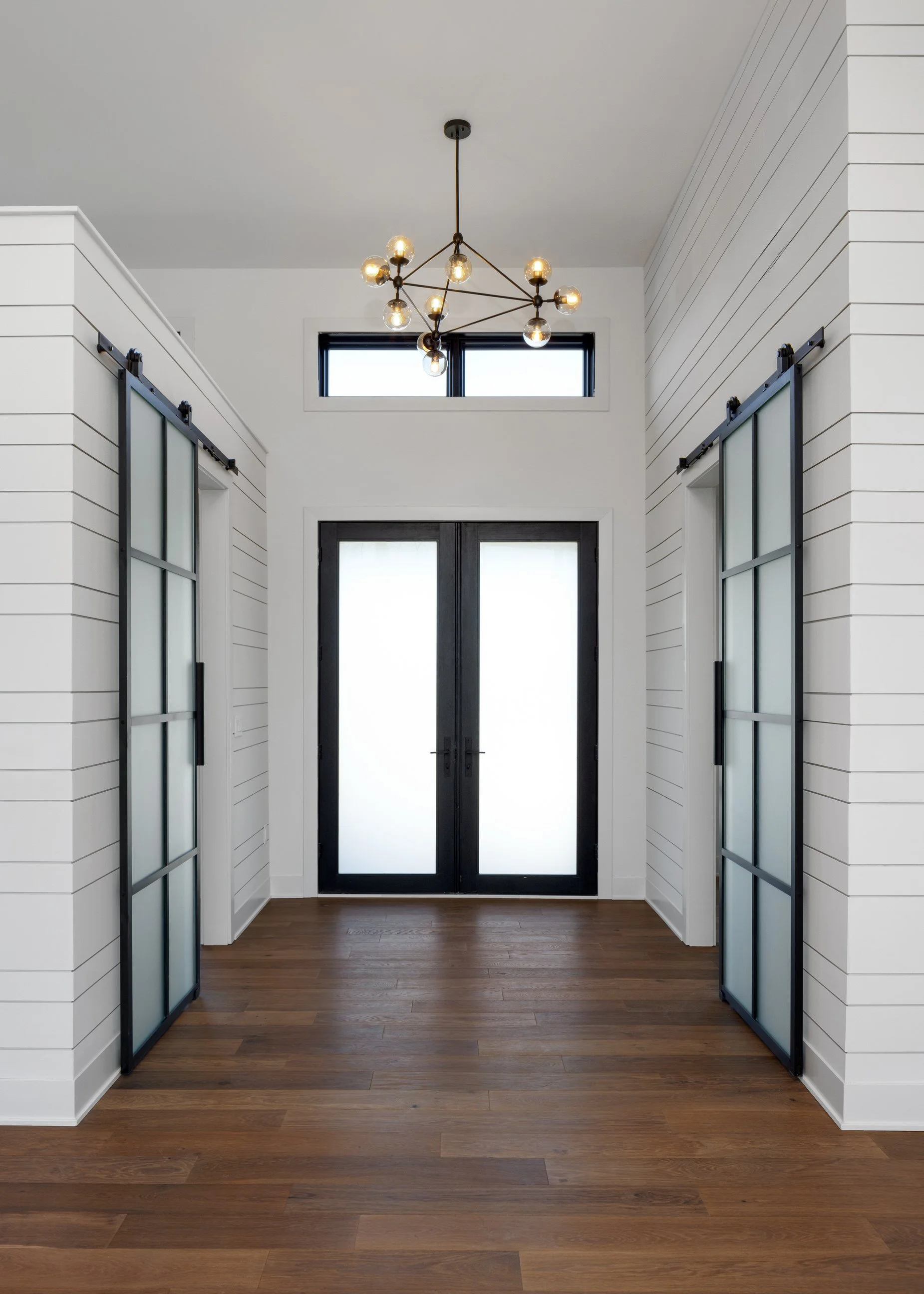 Entryway with hardwood floor, white shiplap walls, black-framed glass double doors, high window above, and modern black chandelier hanging from the ceiling.