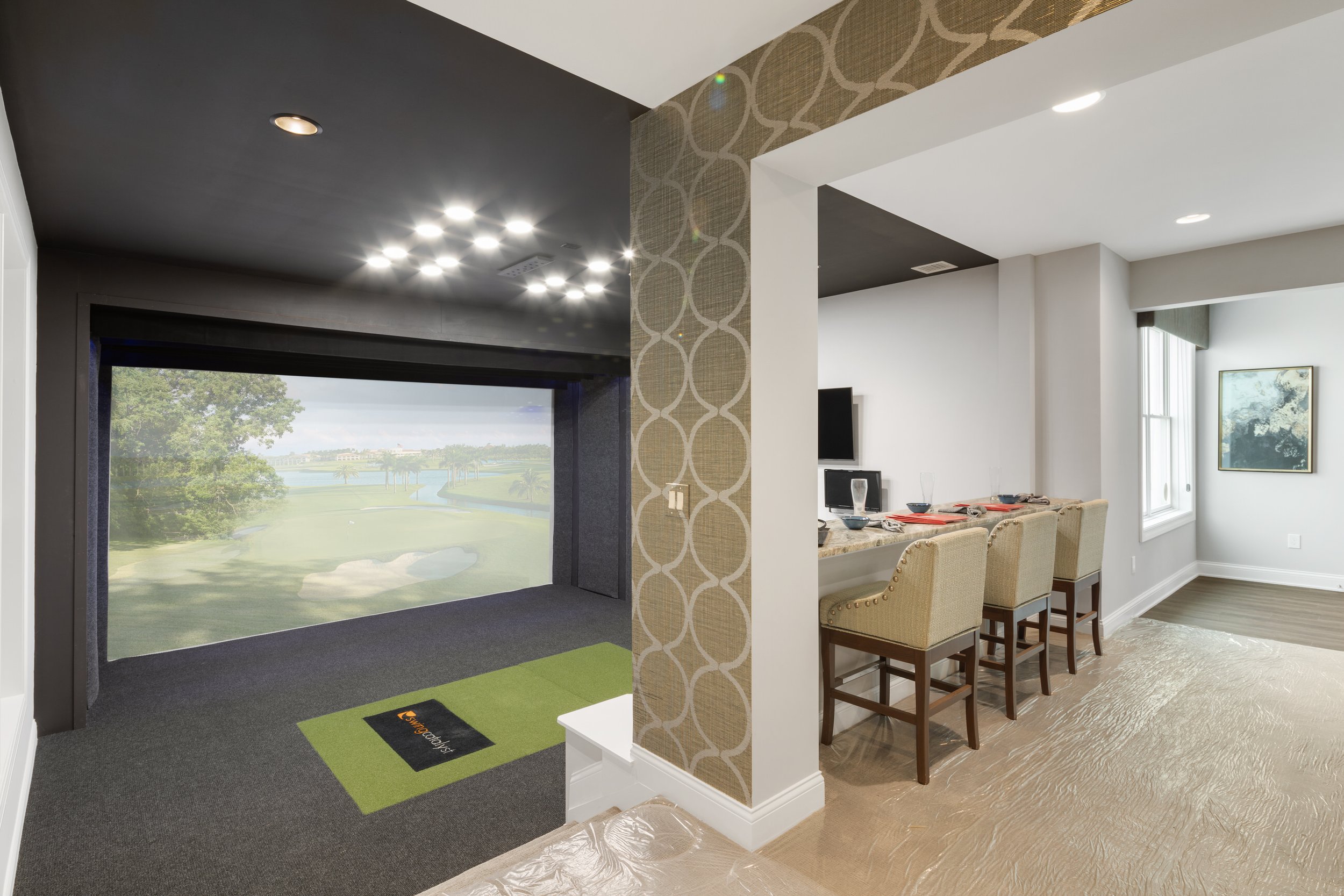 Indoor golf putting area with a large window showing trees and water outside, adjacent to a dining area with a table, chairs, and framed artwork on the wall.