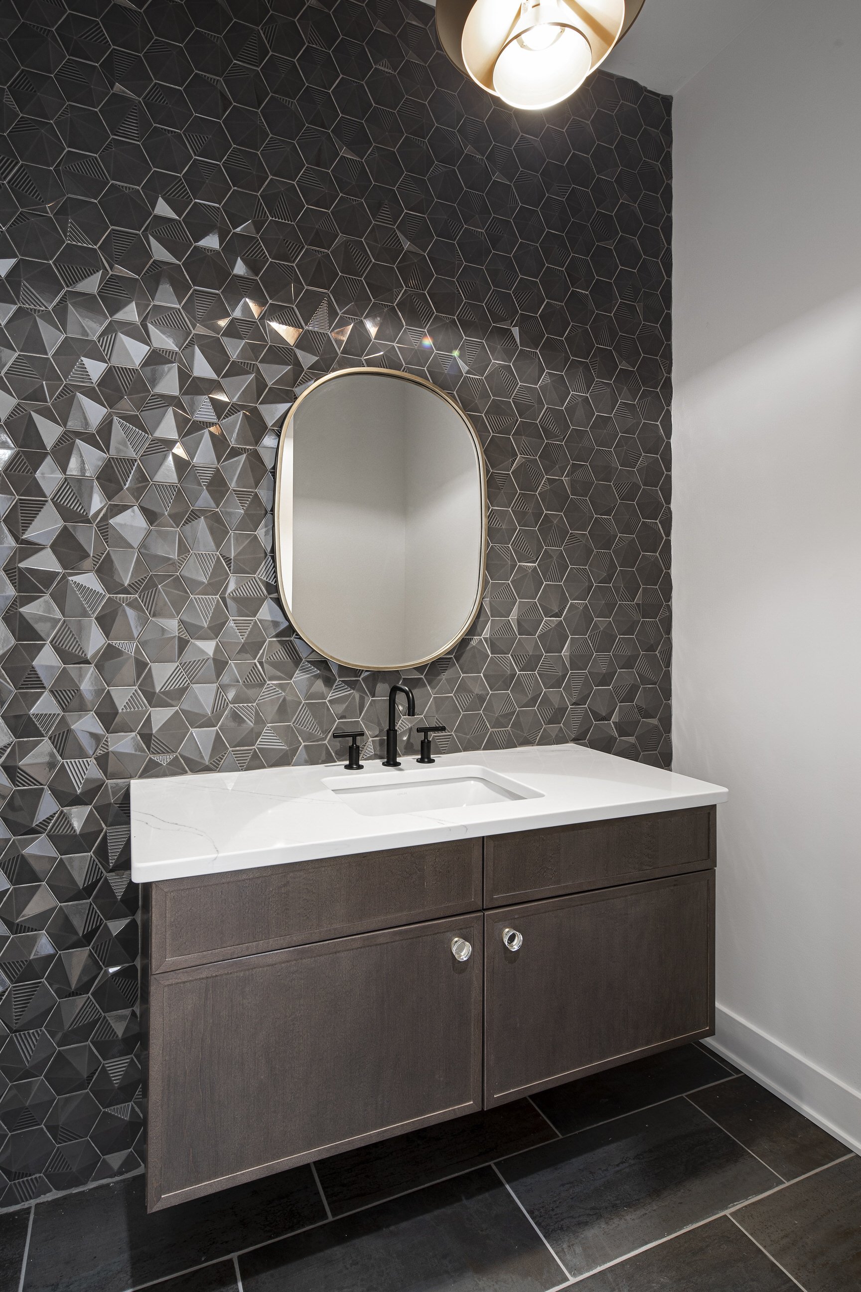 Bathroom vanity with a dark wood cabinet, white marble countertop, black faucet, oval mirror with a gold frame, geometric patterned black tile wall, and black floor tiles.