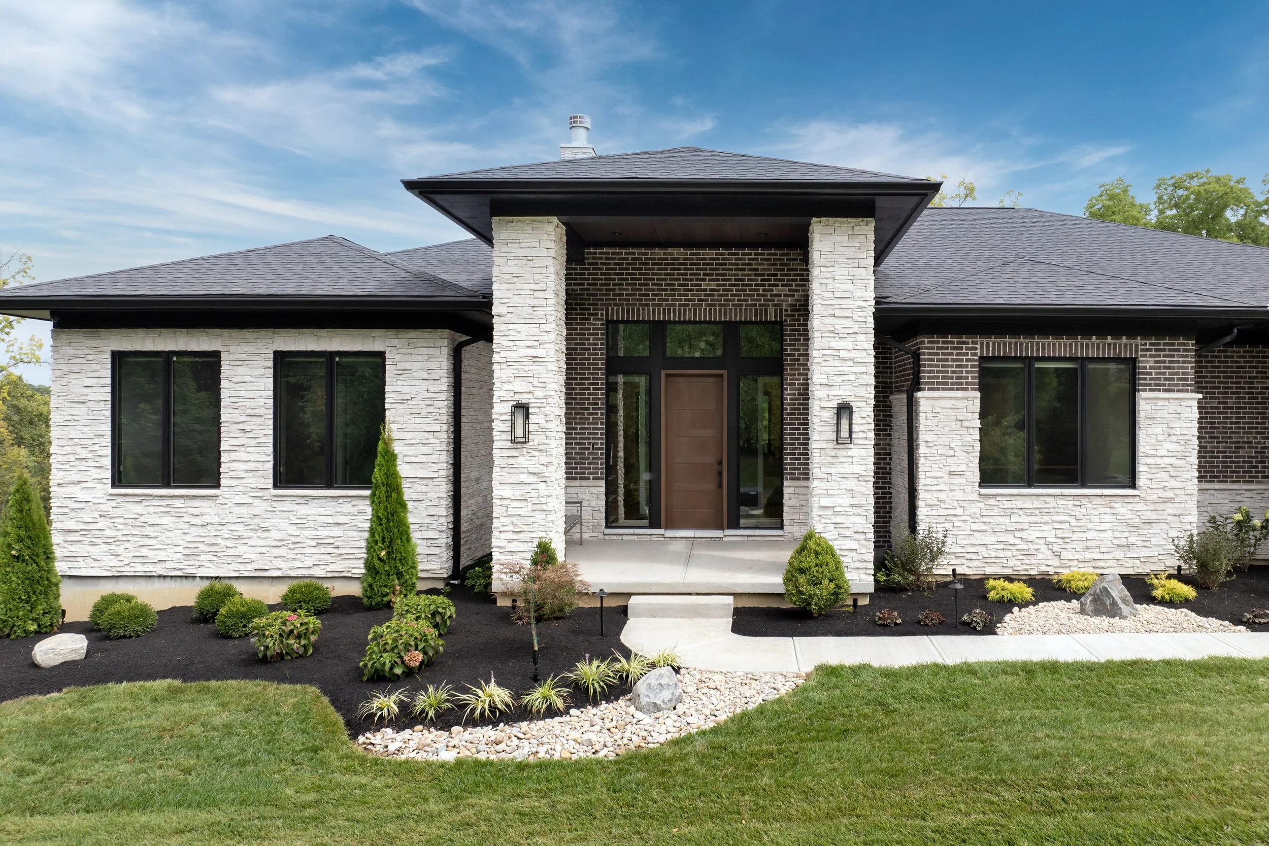 Front view of a custom home with a mix of black and white brick exterior, large windows, and a front porch. There is a landscaped yard with small bushes, rocks, and a green lawn under a partly cloudy blue sky.