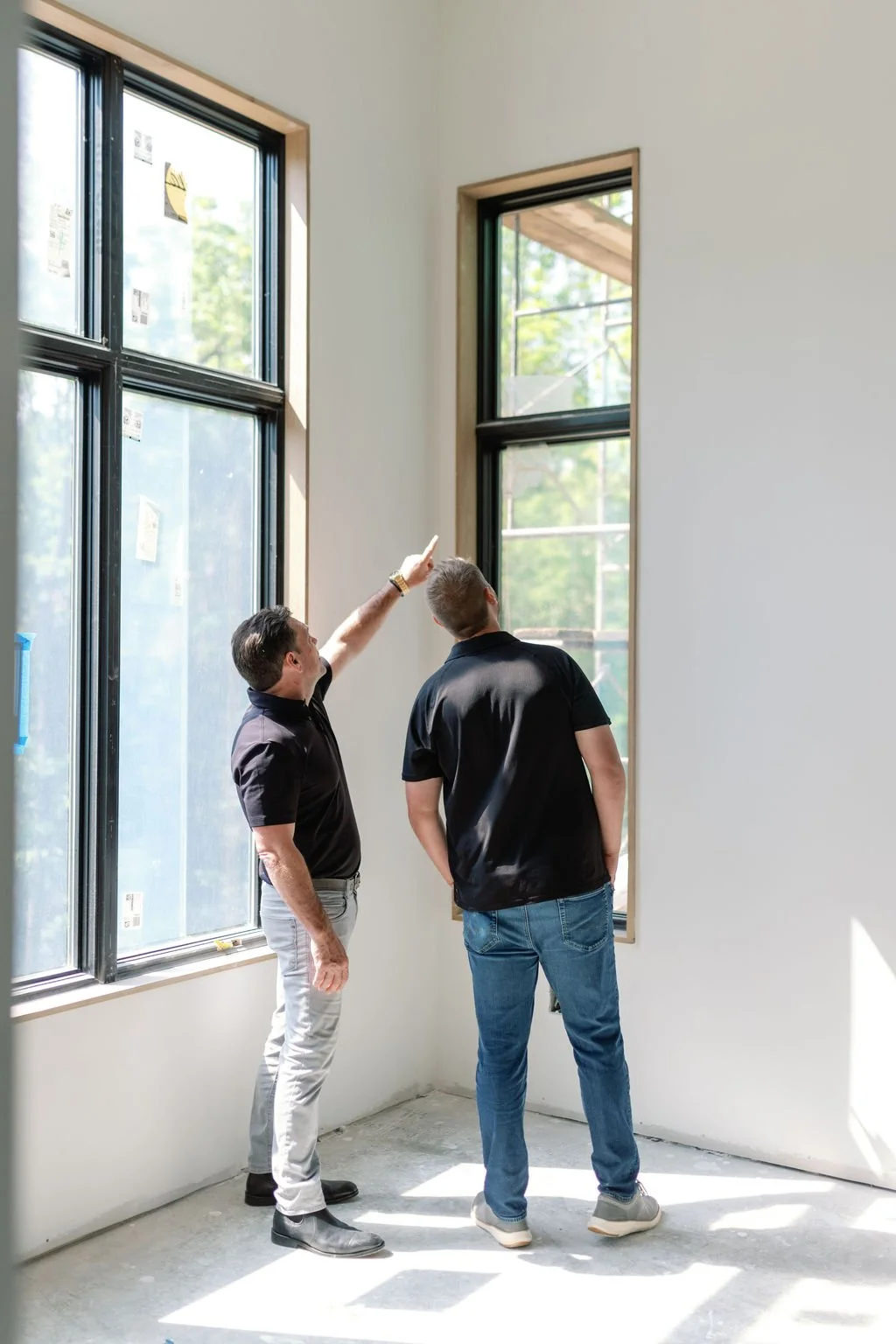 Two men inspecting windows in a room under construction, one pointing at the window while the other looks on.