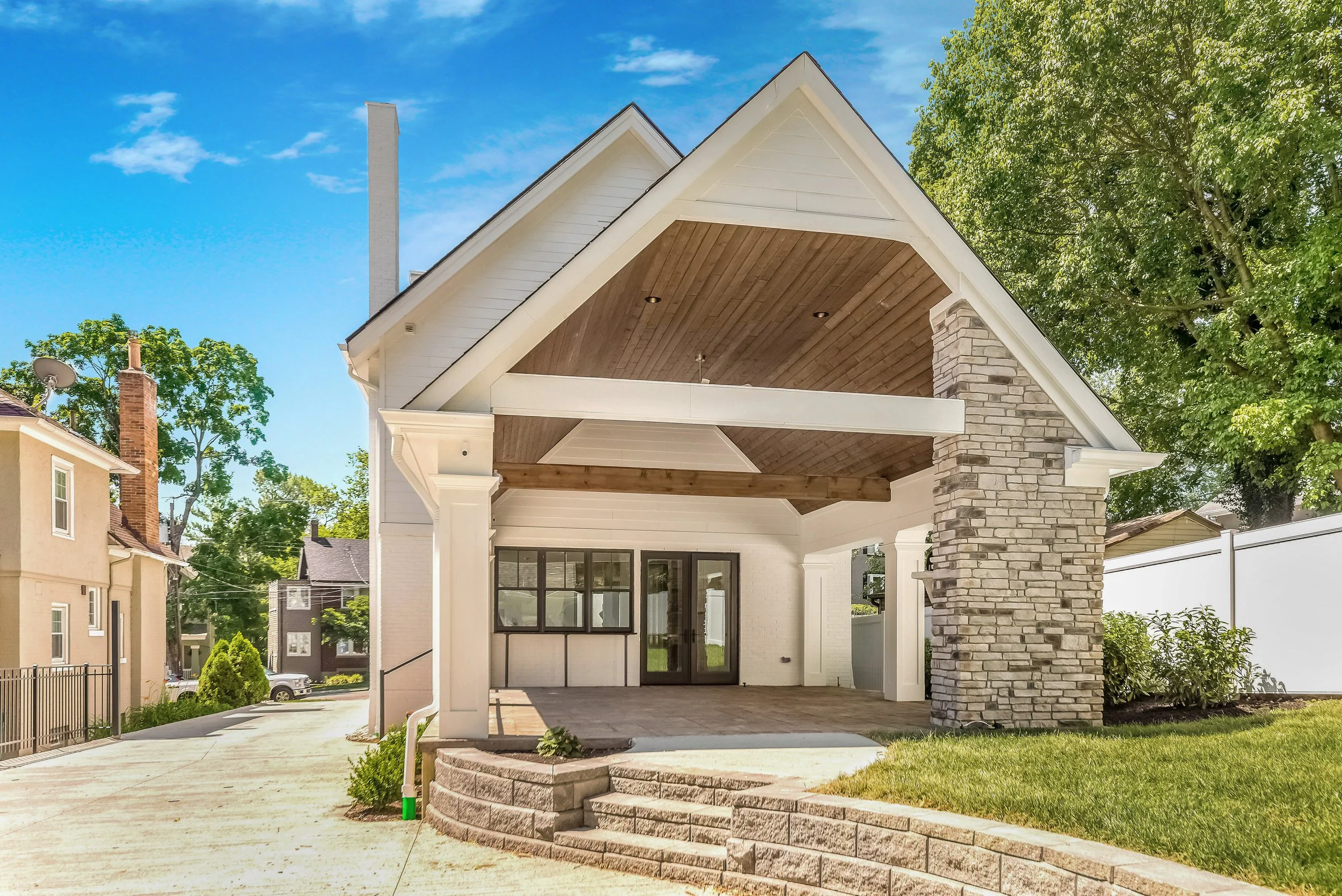 Front view of a house with a covered porch, stone columns, and a gable roof, surrounded by trees and neighboring houses under a blue sky.