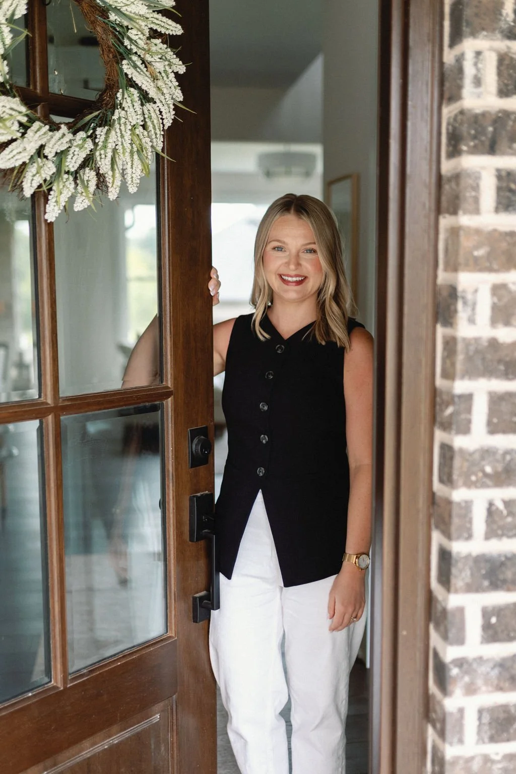 A woman with shoulder-length blonde hair smiling and standing behind a wooden door with glass panels, with a festive wreath hanging on it, in a cozy home interior.