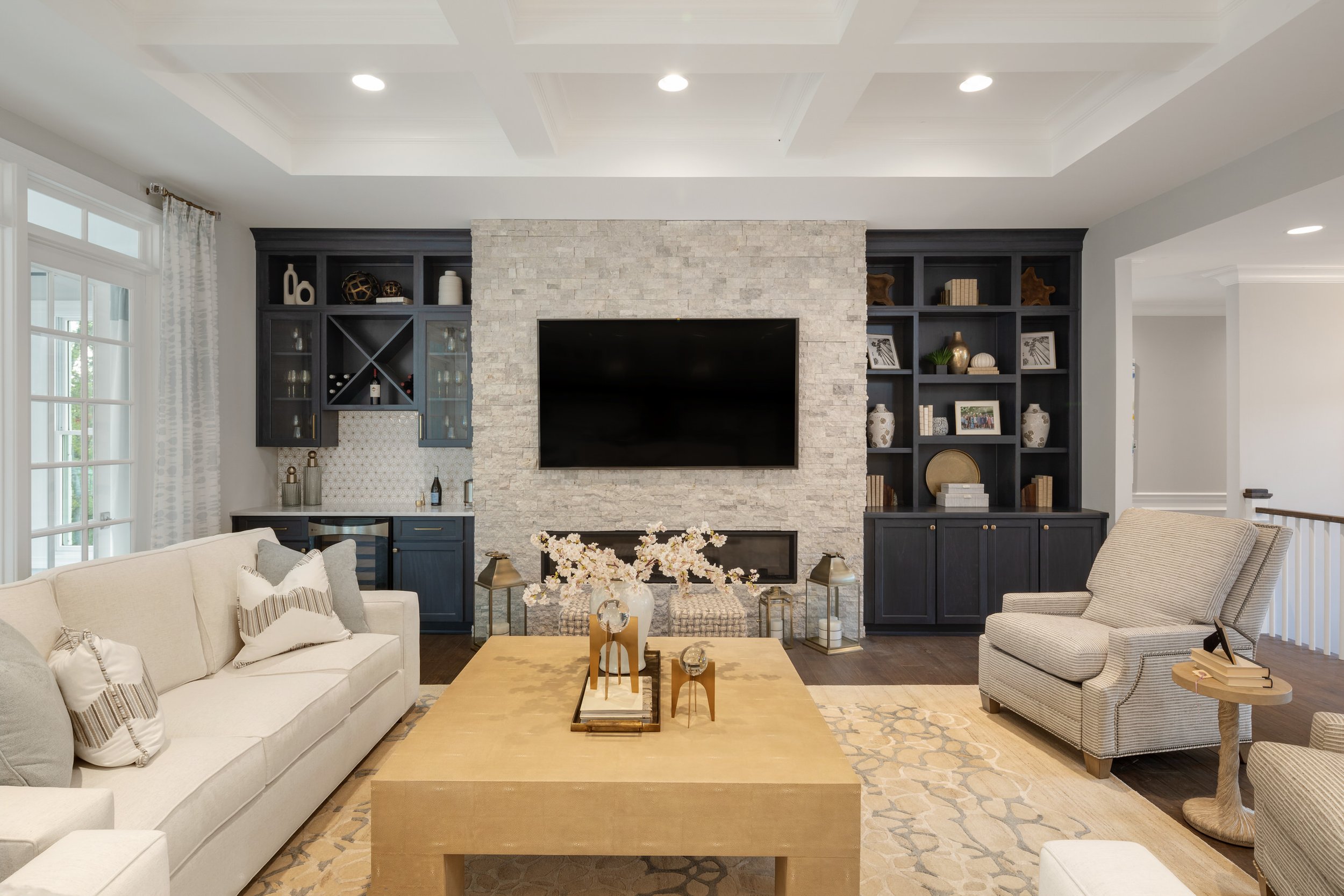 Living room with cream sofa and striped pillows, beige armchair, stone fireplace with TV, navy built-in shelves with decorative items, large window with curtains, wooden coffee table with decor, patterned rug.