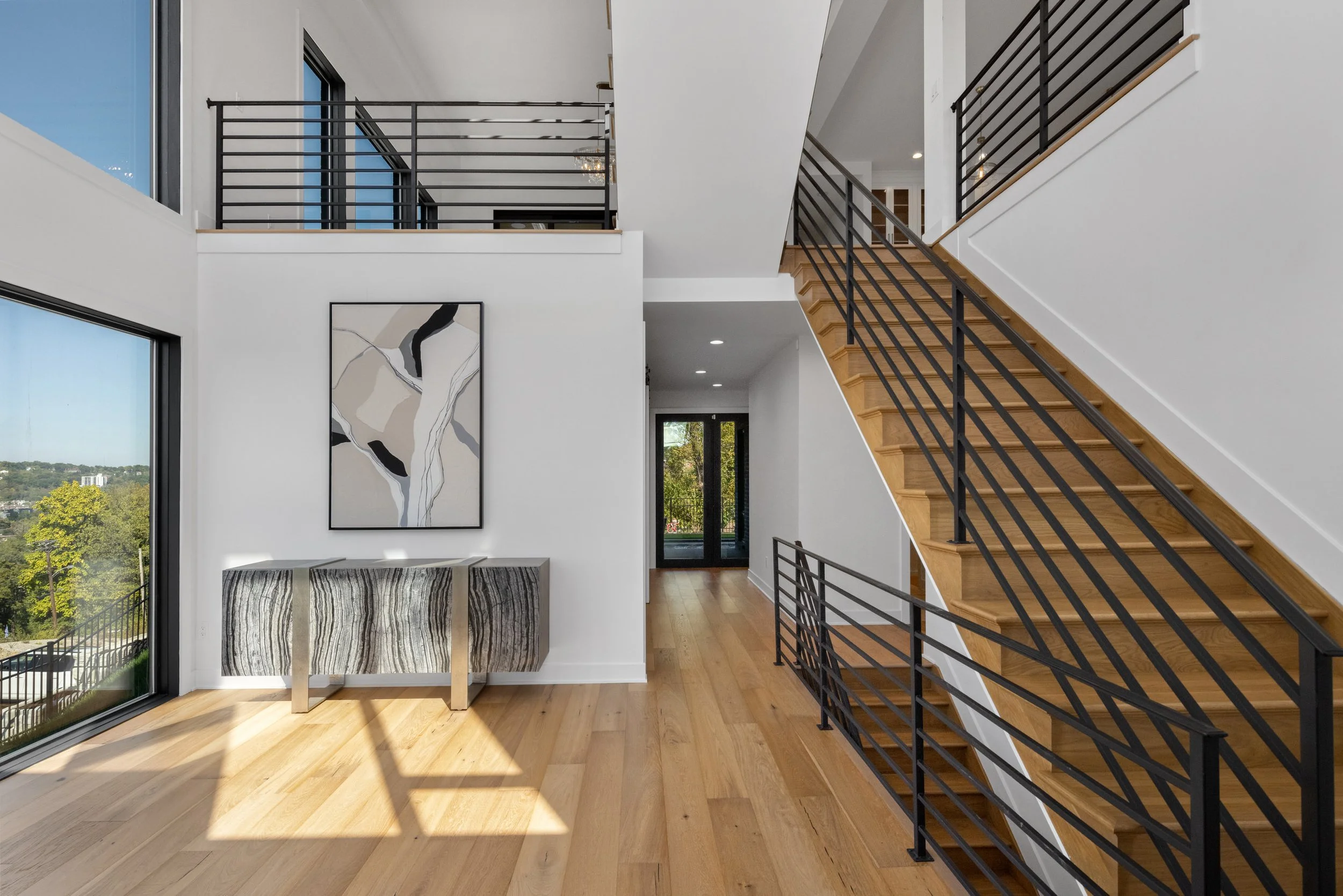 Interior of a modern house with large windows, a staircase with black metal railings, and light-colored hardwood floors. There's abstract artwork on the wall and a console table beneath it. Natural light floods the space, showing a view of trees outside.