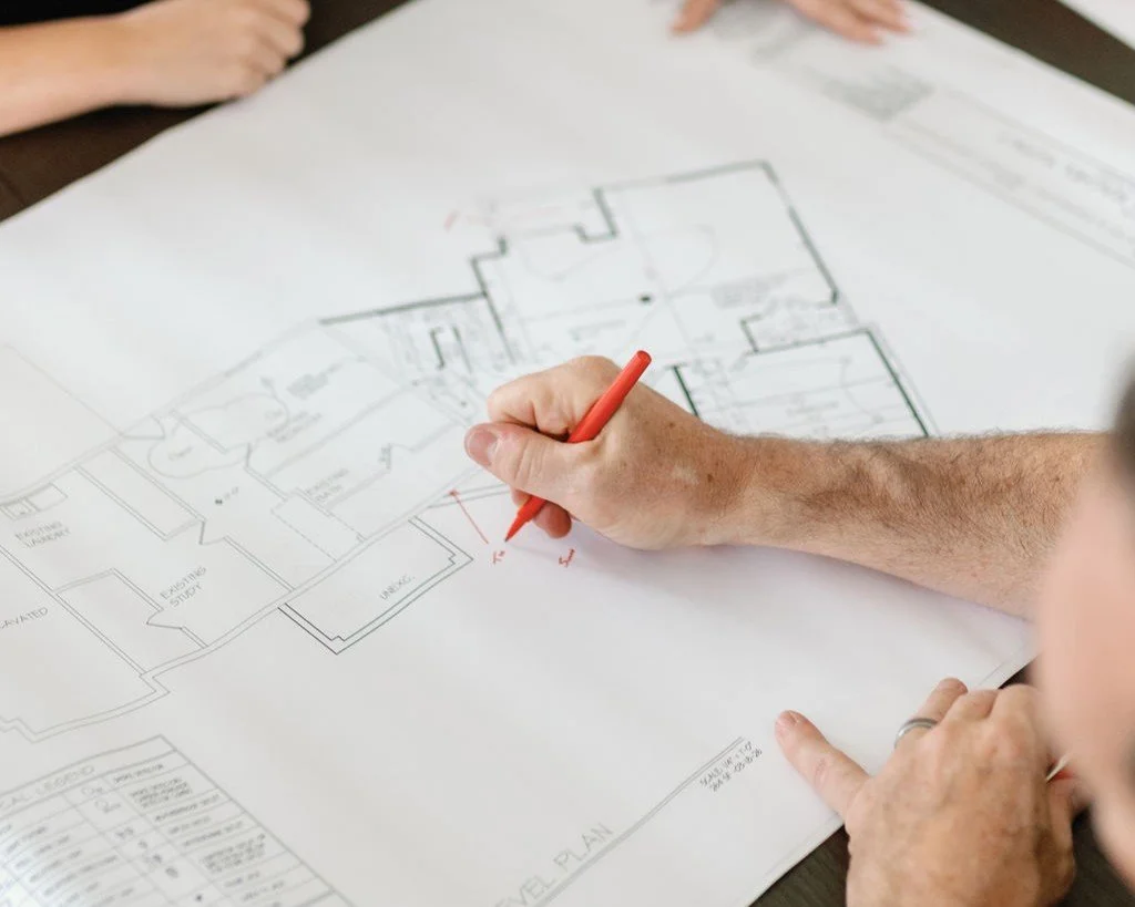 A person drawing on an architectural blueprint with a red marker, with other hands and a part of a person's face visible in the background.