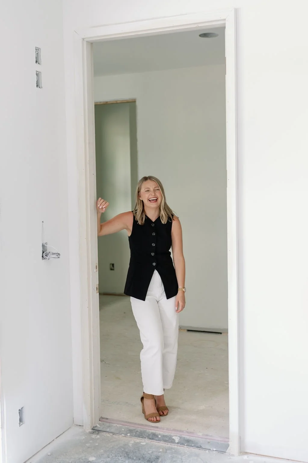A woman laughing joyfully in a room under construction, standing in a doorway, wearing a black sleeveless top and white pants.