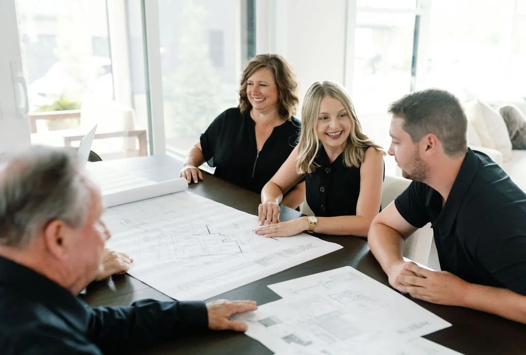 Redknot Custom Home team gathered around a table looking at architectural plans and blueprints in a bright, modern room with large windows.