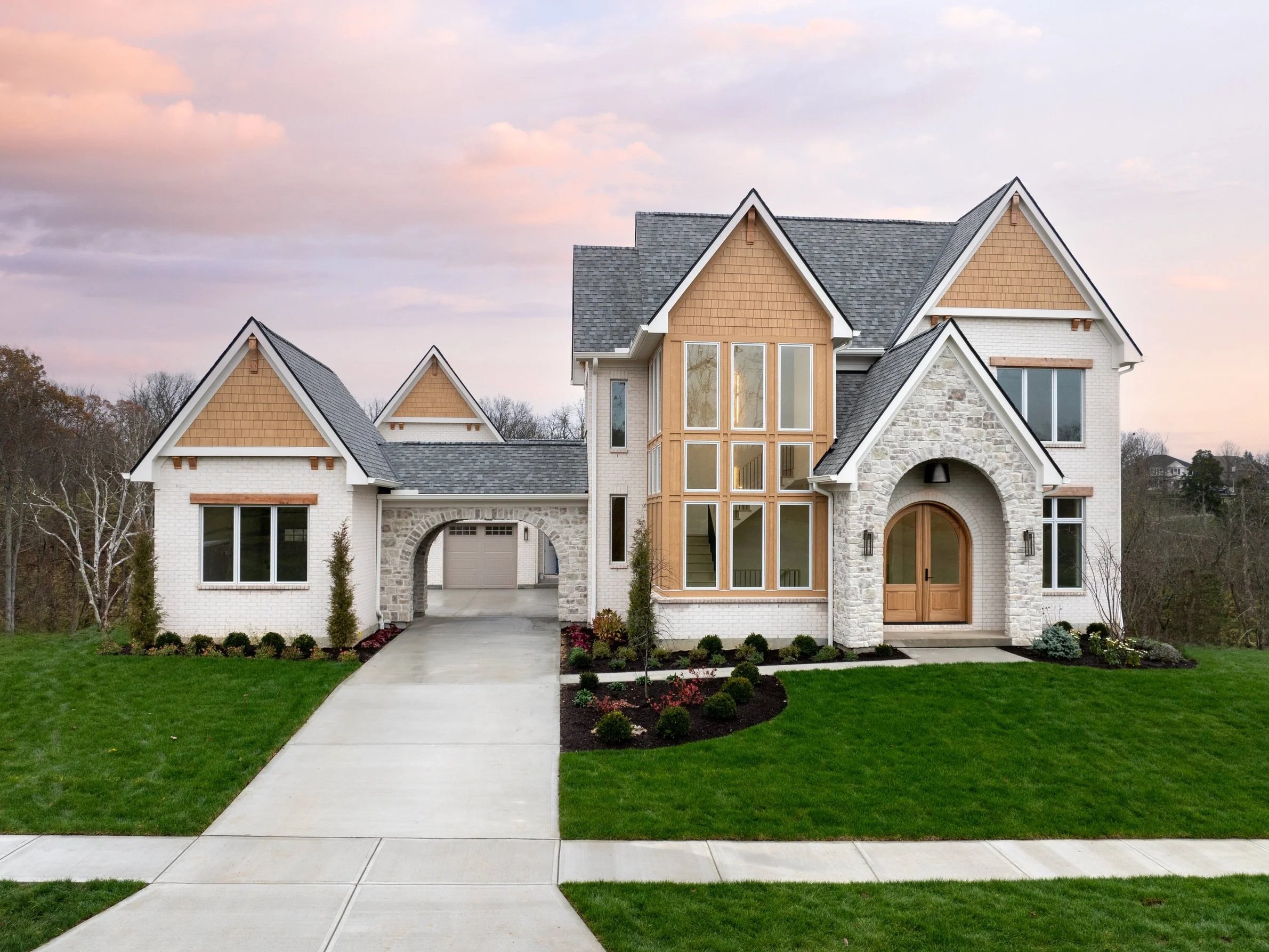 A modern two-story house with a combination of brick, stone, and wood exterior, multiple peaked roofs, large windows, a central arched entryway, and a paved walkway through a well-manicured lawn.