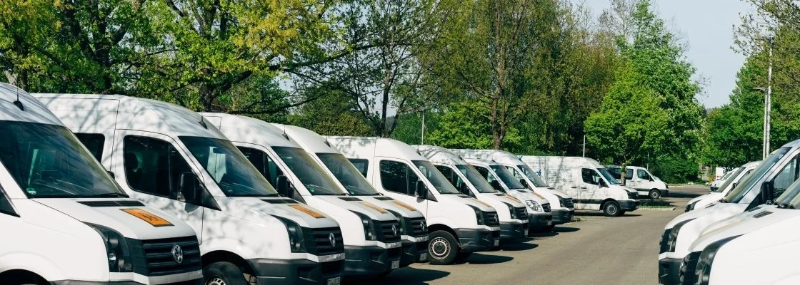 A row of white commercial delivery vans parked in a lot with trees in the background.