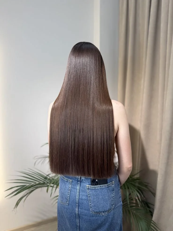Mujer con cabello largo, liso, brillante y castaño oscuro, vista trasera, usando un pantalón de mezclilla, en un ambiente interior con plantas y cortinas beige.