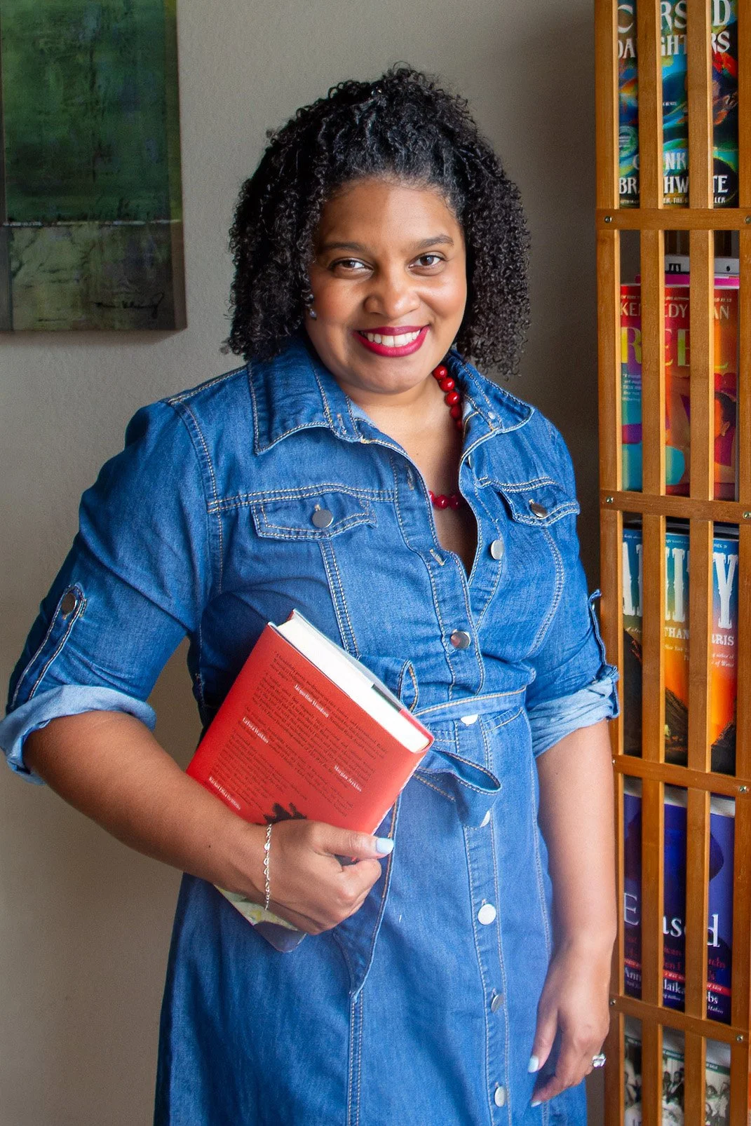 Kami Tei wearing a denim dress with rolled-up sleeves, holding a red book, standing in front of a bookshelf with colorful books.