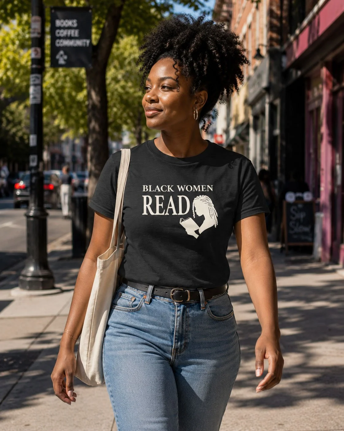 A woman wearing a black T-shirt with the words 'Black Women Read' and an illustration of a woman reading a book, standing on a city sidewalk with trees and buildings in the background.