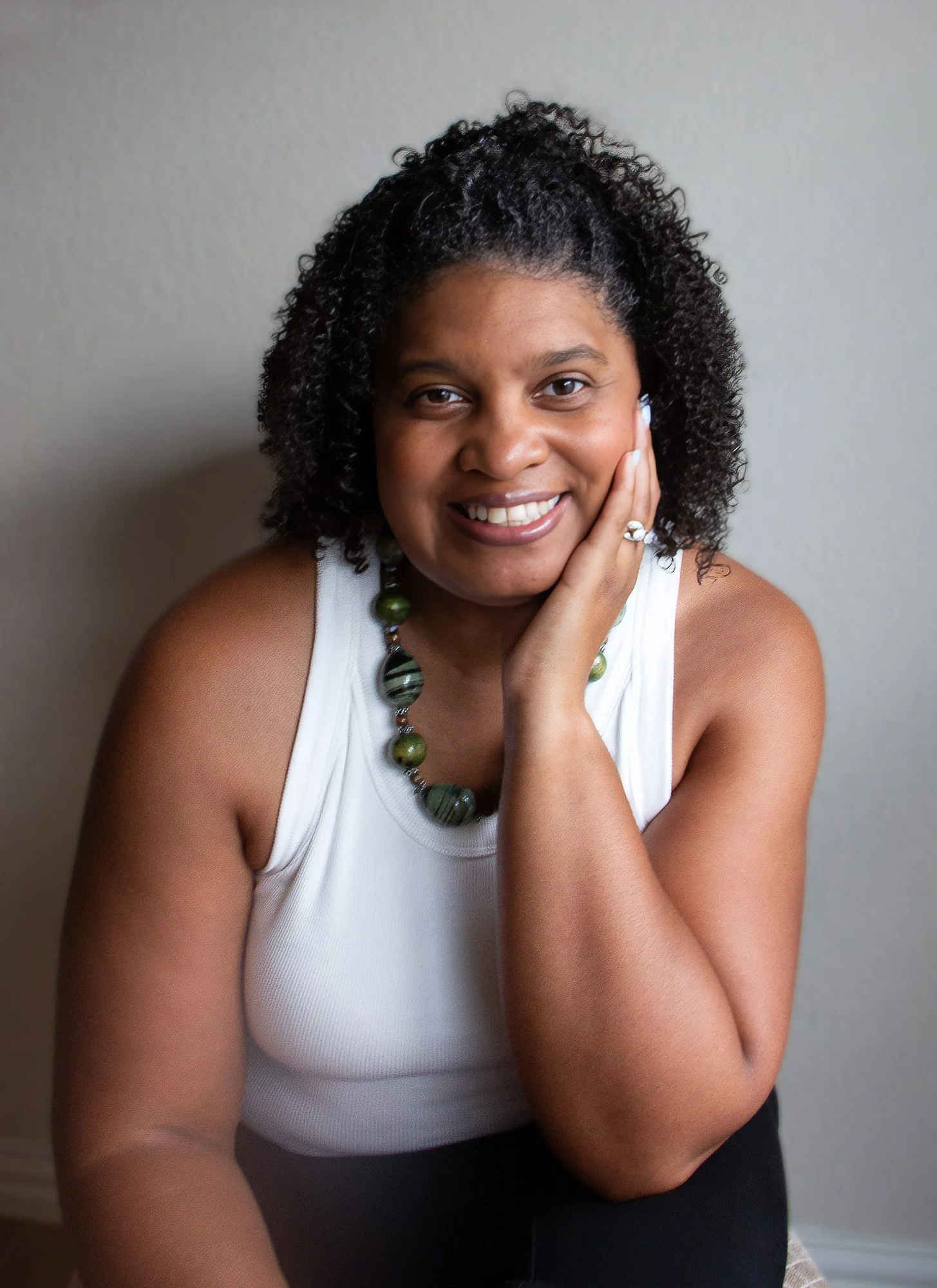 Kami Tei smiling with her hand on her face, wearing a white sleeveless top and a necklace with large green and black beads.