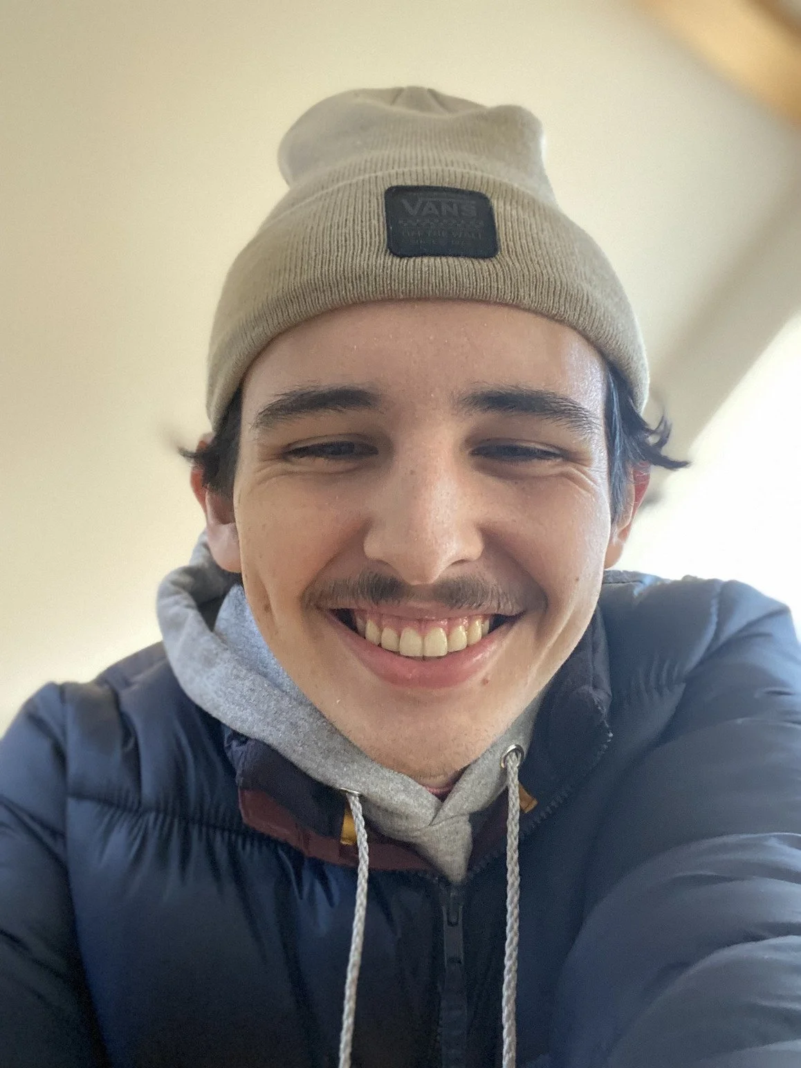 A young man wearing a beige beanie, a gray hoodie, and a black jacket, smiling and taking a selfie indoors.