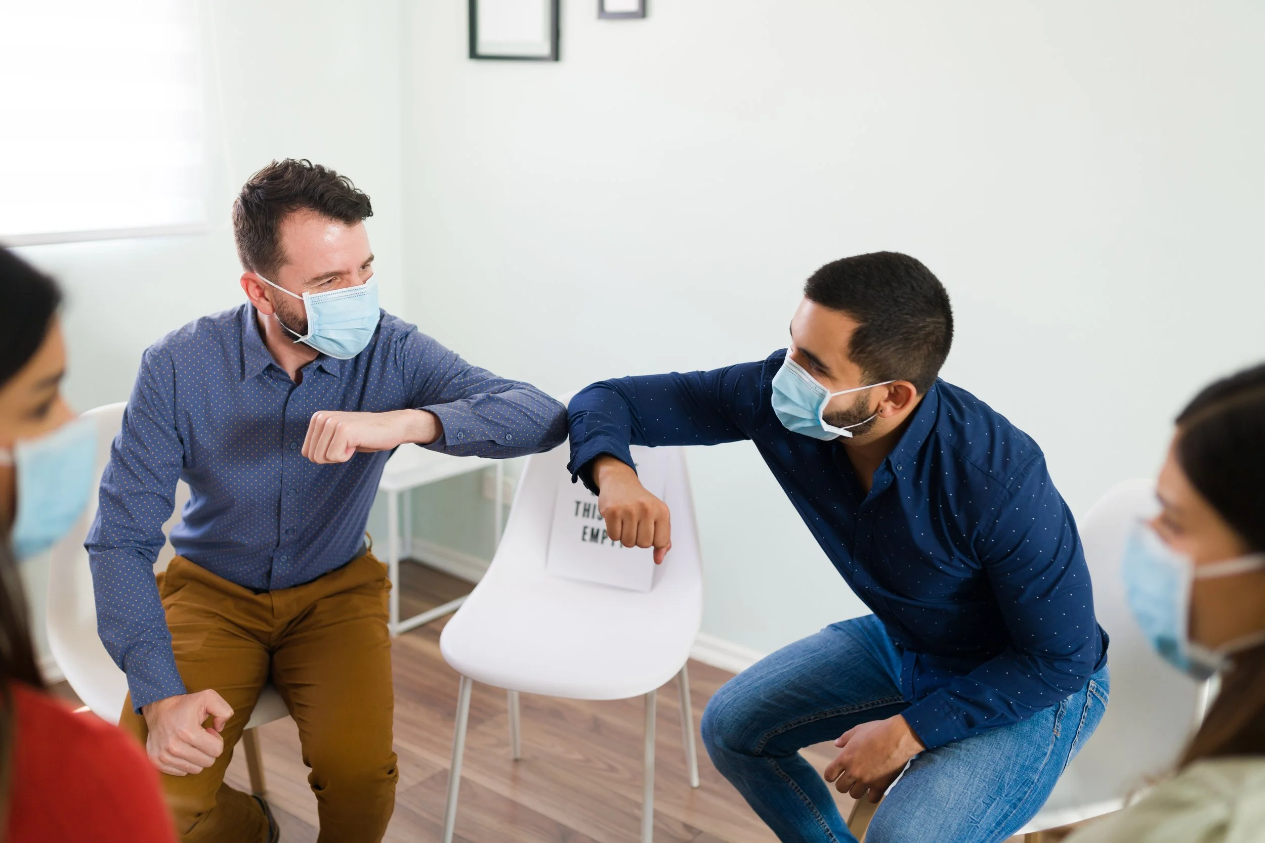 People wearing face masks in a meeting, engaging in a fist bump for greeting or camaraderie in a discussion.