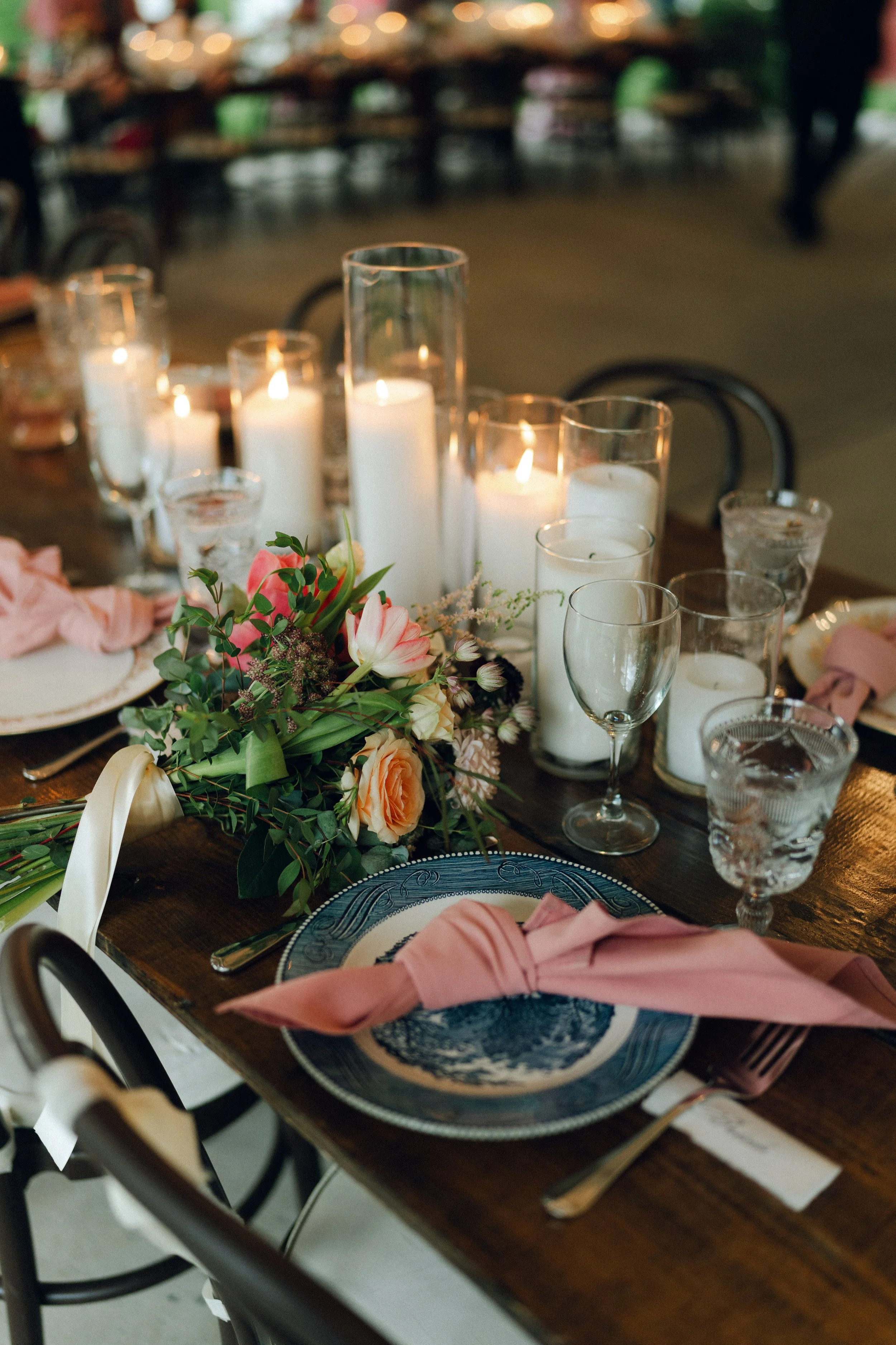 A decorated table set for a special occasion, featuring a floral centerpiece, lit candles in various glass holders, elegant china, pink napkins, and glassware.