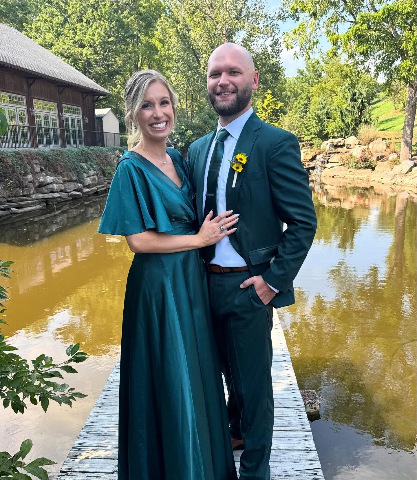 A smiling woman in a teal dress and a man in a dark suit with a yellow sunflower boutonniere, standing on a wooden dock by a pond with trees in the background.