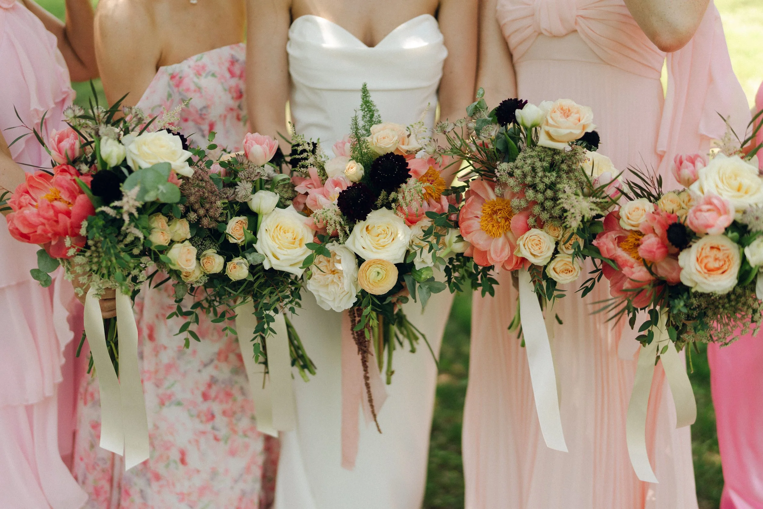 People holding floral arrangements at an outdoor event, likely a wedding, with pastel pink, peach, and white flowers.