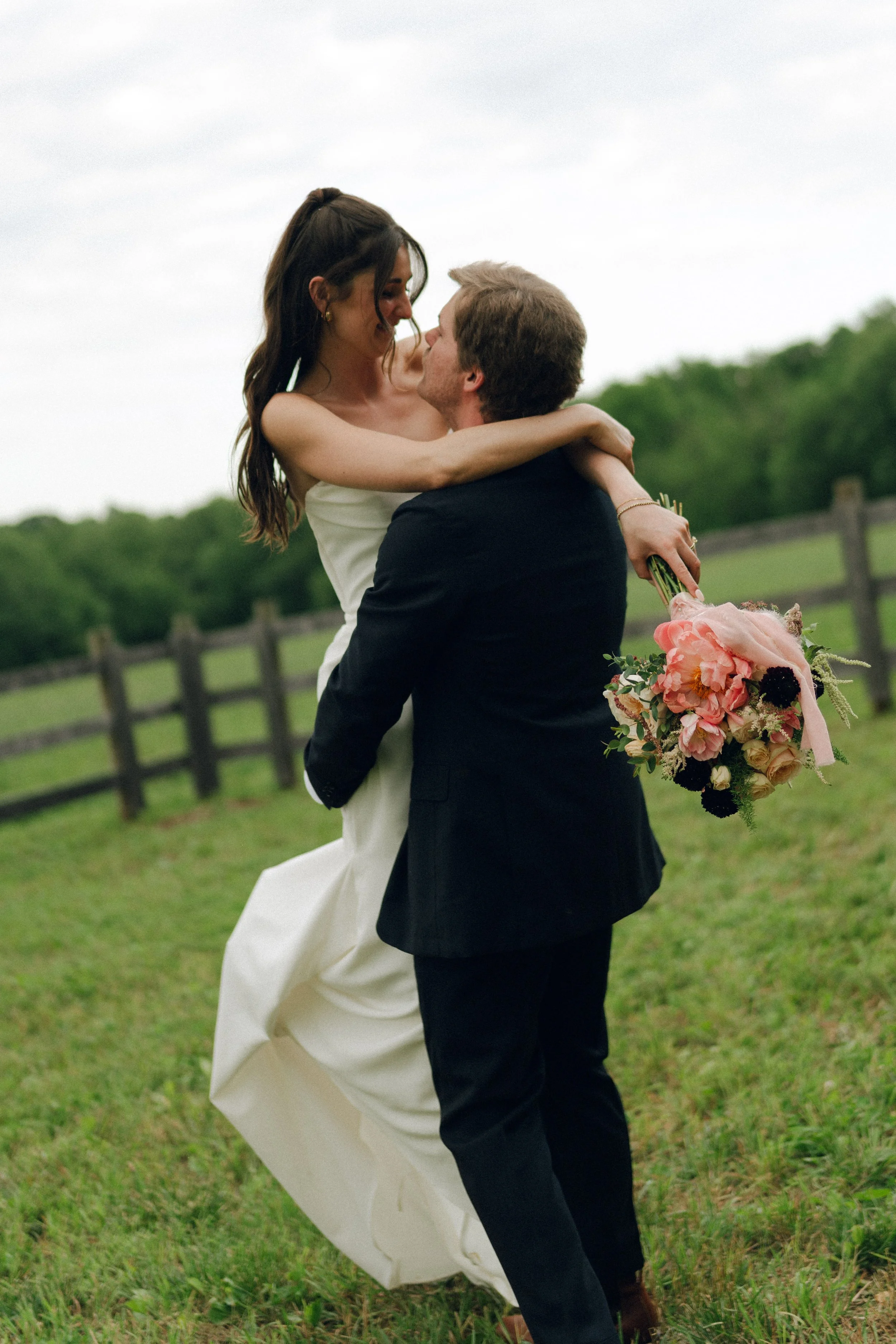 A couple is embracing outdoors on a grassy field, the woman wearing a white dress and the man in a dark suit, holding a bouquet of flowers.