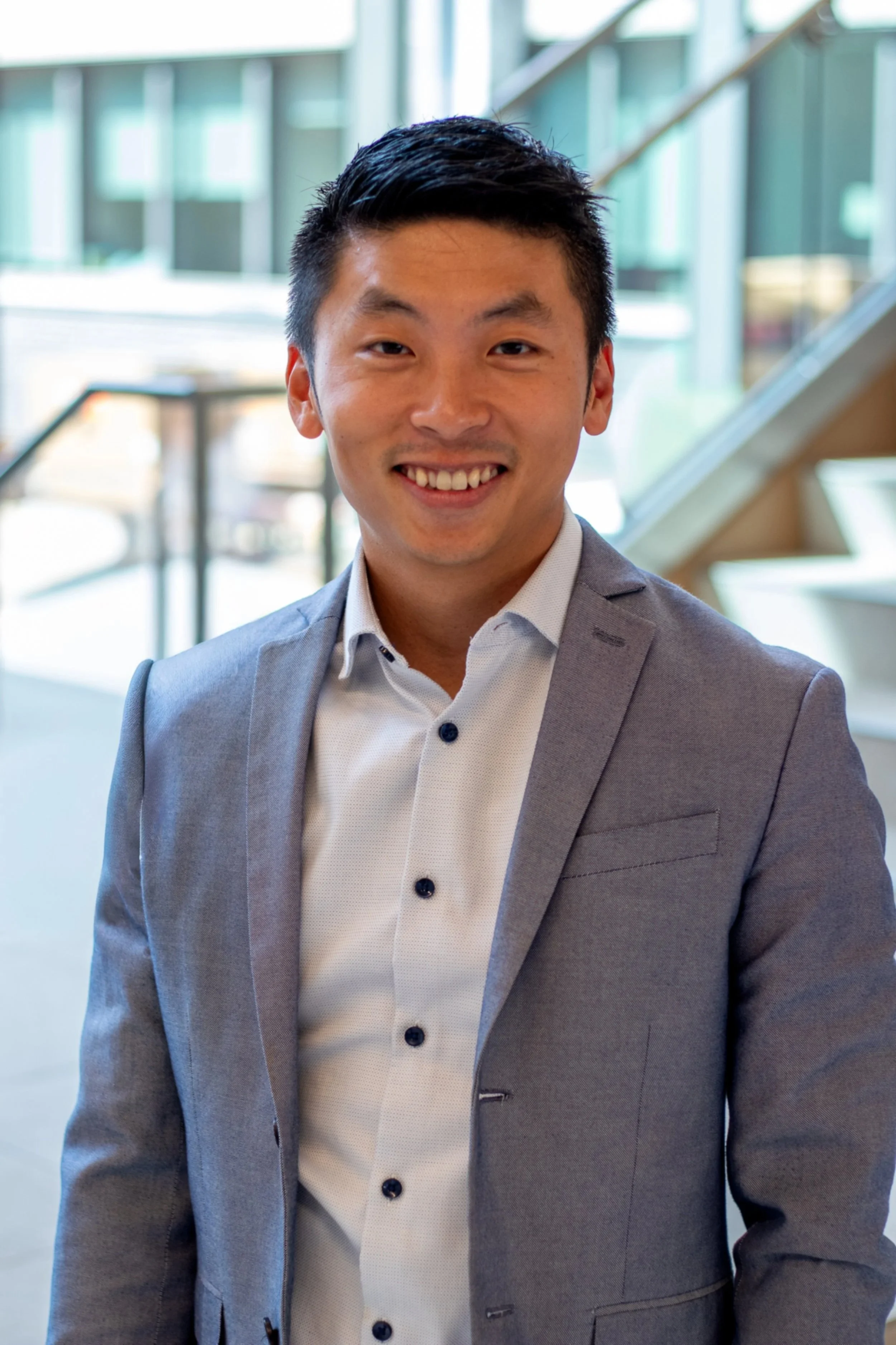 A young man in a light gray suit jacket and white shirt smiling inside a modern building with glass walls and a staircase in the background.