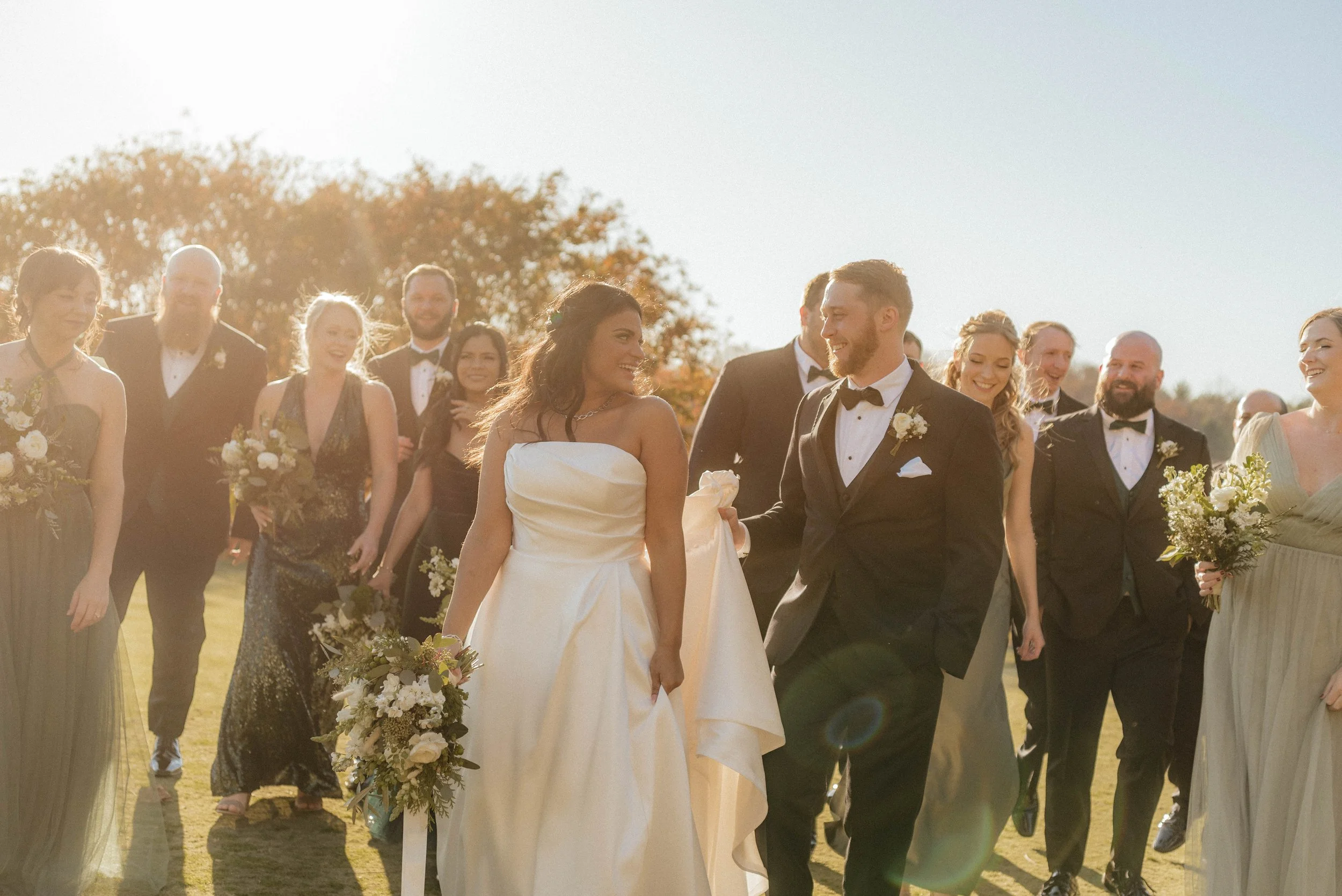 A bride and groom holding hands and walking during their outdoor wedding ceremony, surrounded by smiling bridesmaids and groomsmen in formal attire on a sunny day.