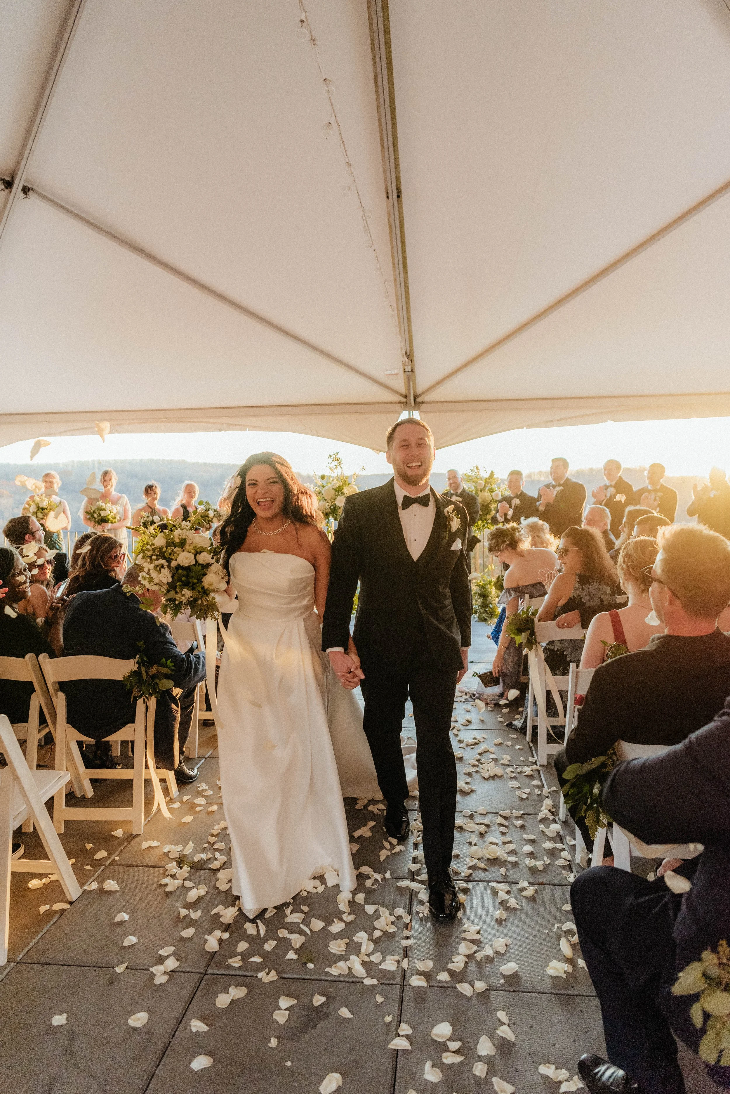 A bride and groom walking down an aisle under a canopy during a wedding ceremony. The bride is wearing a strapless white dress and holding a large bouquet, and the groom is wearing a black tuxedo with a bow tie. The aisle is decorated with scattered flower petals, and guests are seated on either side, watching and applauding.