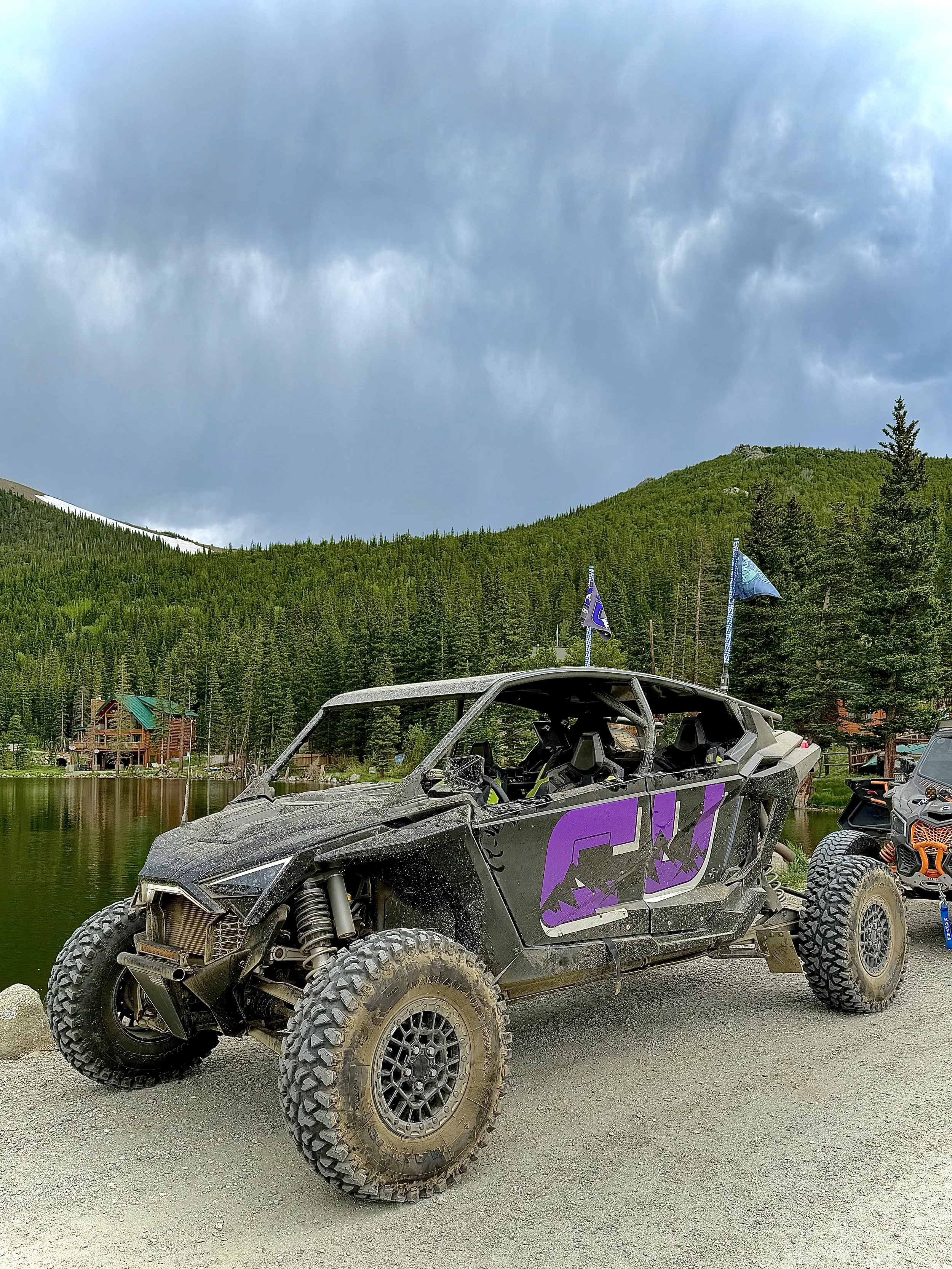 A black off-road vehicle with purple graphics, large tires, and a partially exposed interior parked near a lake with a forested hill and overcast sky in the background.