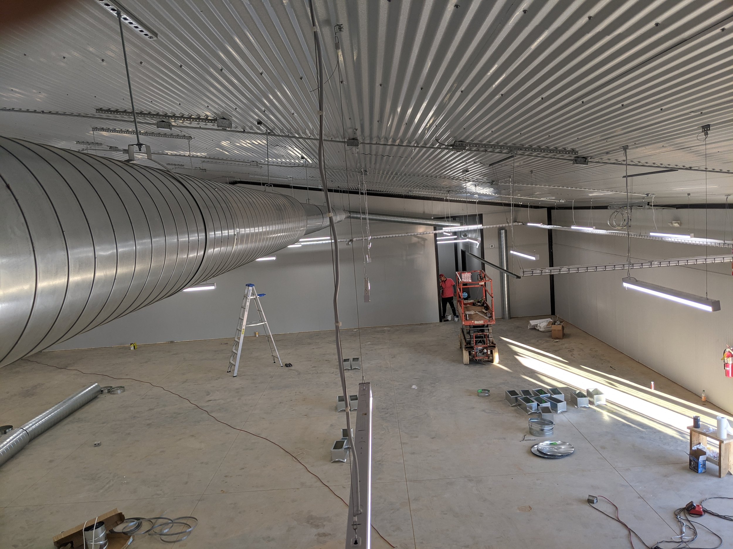 Construction workers installing large ventilation ductwork in an industrial space with a corrugated metal ceiling and hanging lighting fixtures.