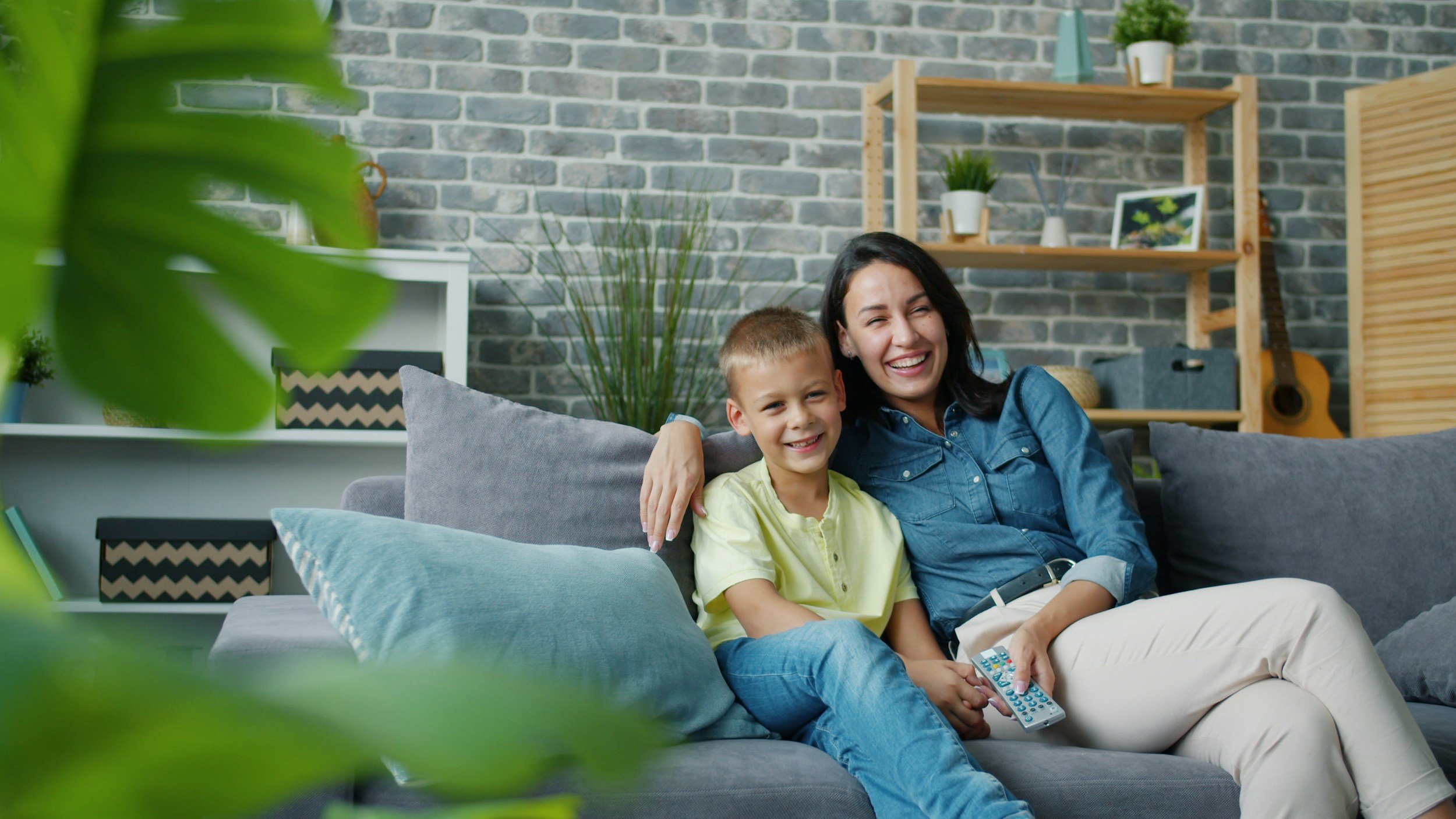 A woman and a young boy sitting on a gray sofa, smiling and enjoying each other's company in a cozy living room with a brick wall, plants, and shelves in the background.