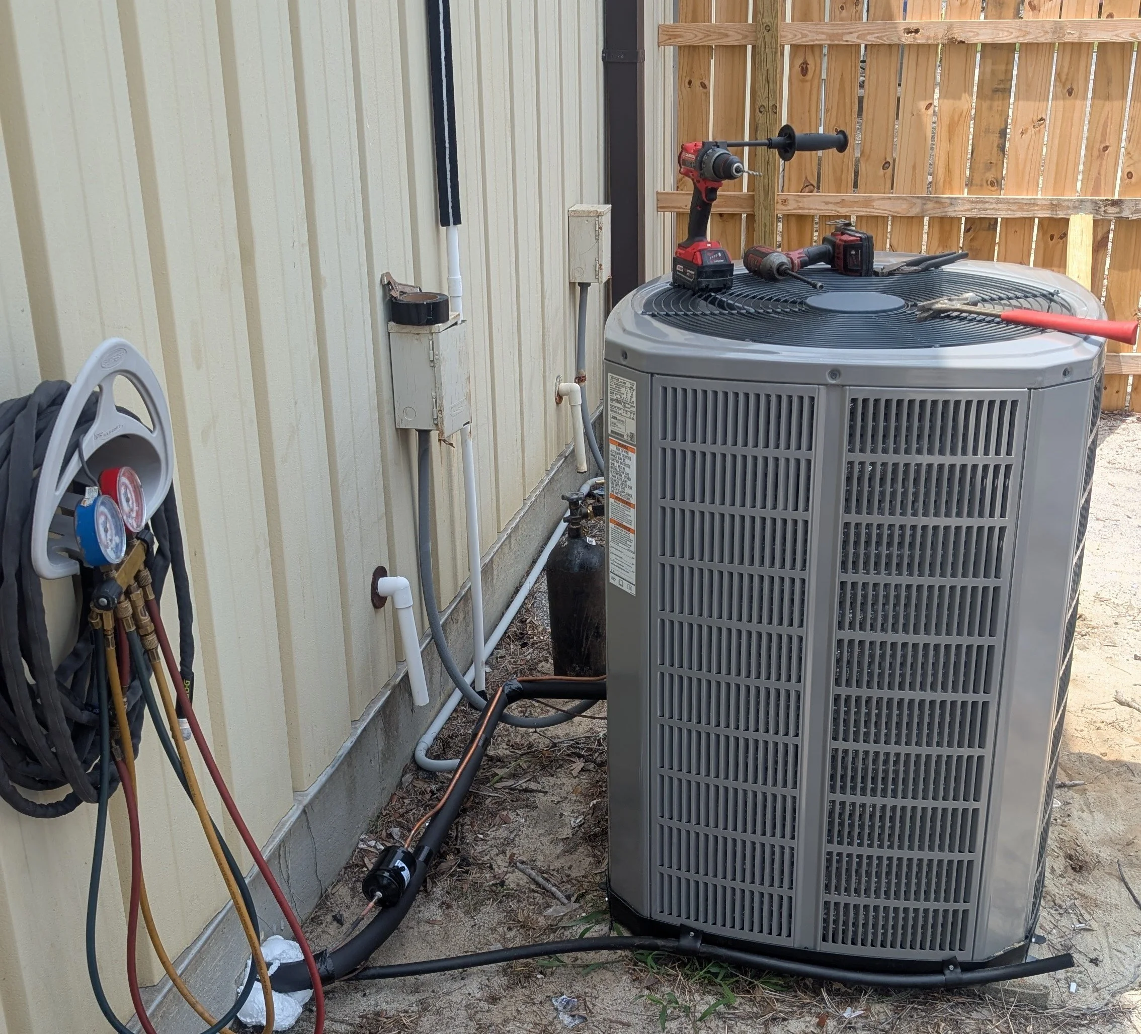 Outdoor HVAC unit with tools and hoses placed on top and nearby, adjacent to a yellow building with vertical siding and a wooden fence in the background.
