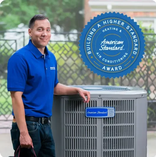 A man in a blue polo shirt standing next to an American Standard air conditioning unit with a badge indicating it has built a higher standard award for heating and air conditioning.