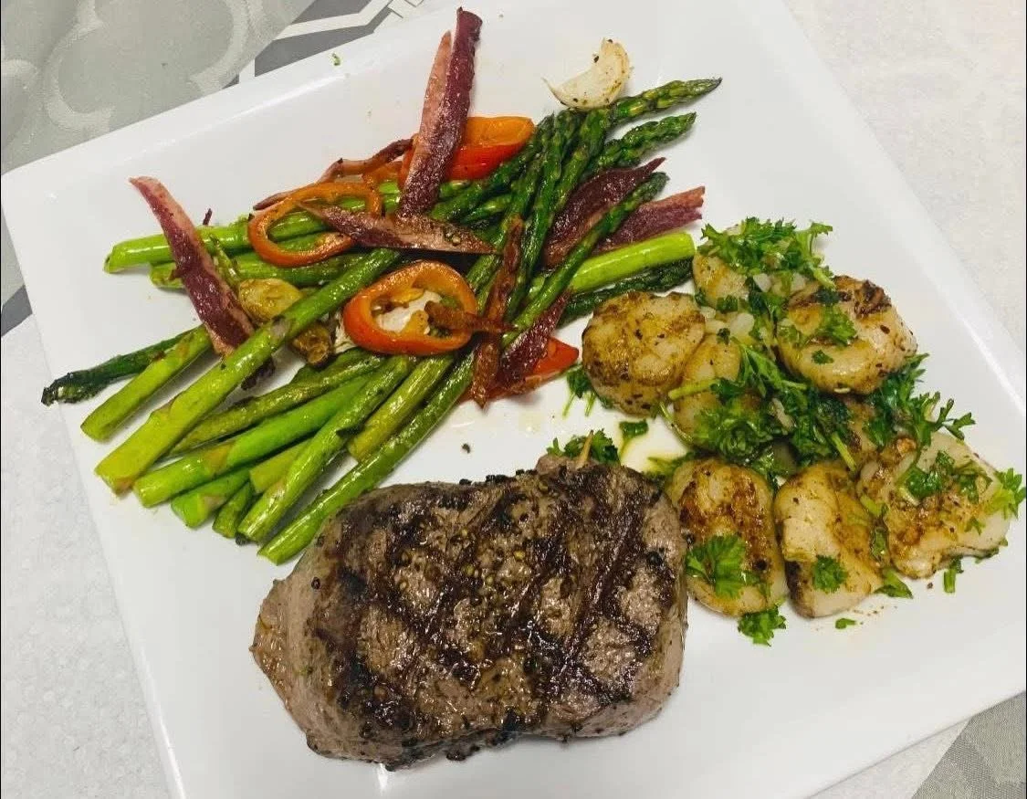 Grilled steak, roasted scallops with herbs, sautéed asparagus, and roasted vegetables on a white square plate.