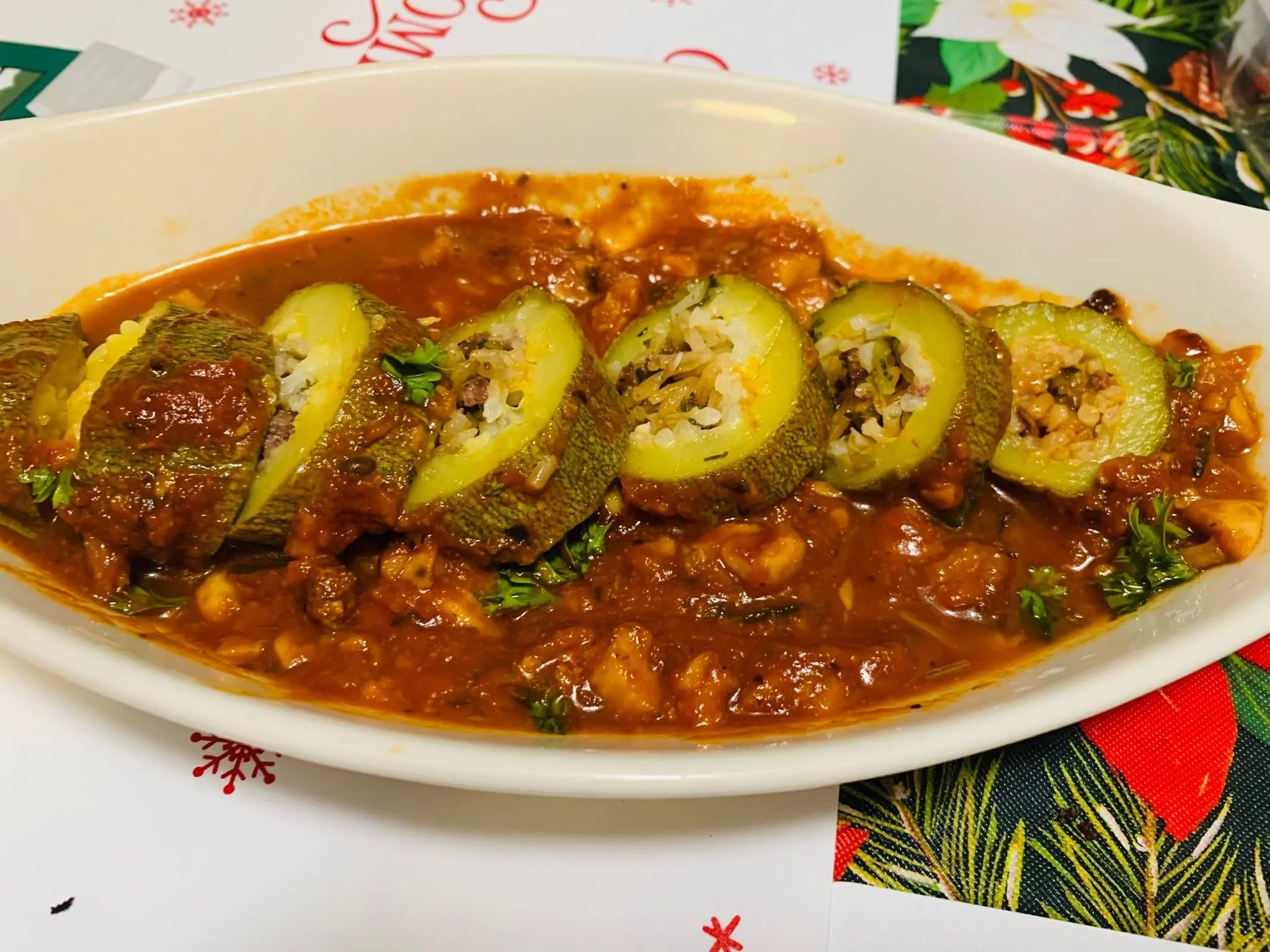 Stuffed zucchini boats topped with tomato sauce and herbs on a white dish, with a colorful holiday-themed tablecloth and wrapped gifts in the background.