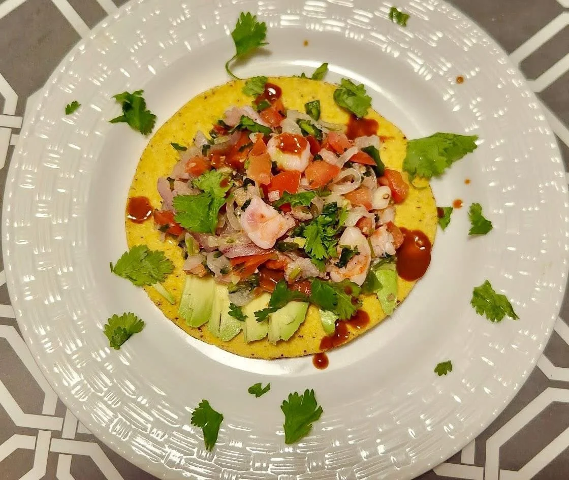A plate of ceviche garnished with cilantro and avocado slices, served on a corn tortilla with sauce drizzled on top.