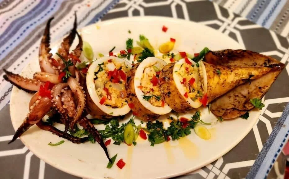 Grilled stuffed calamari, topped with chopped red peppers and herbs, served with whole grilled squid tentacles on a white oval plate, garnished with parsley and sliced green onions.