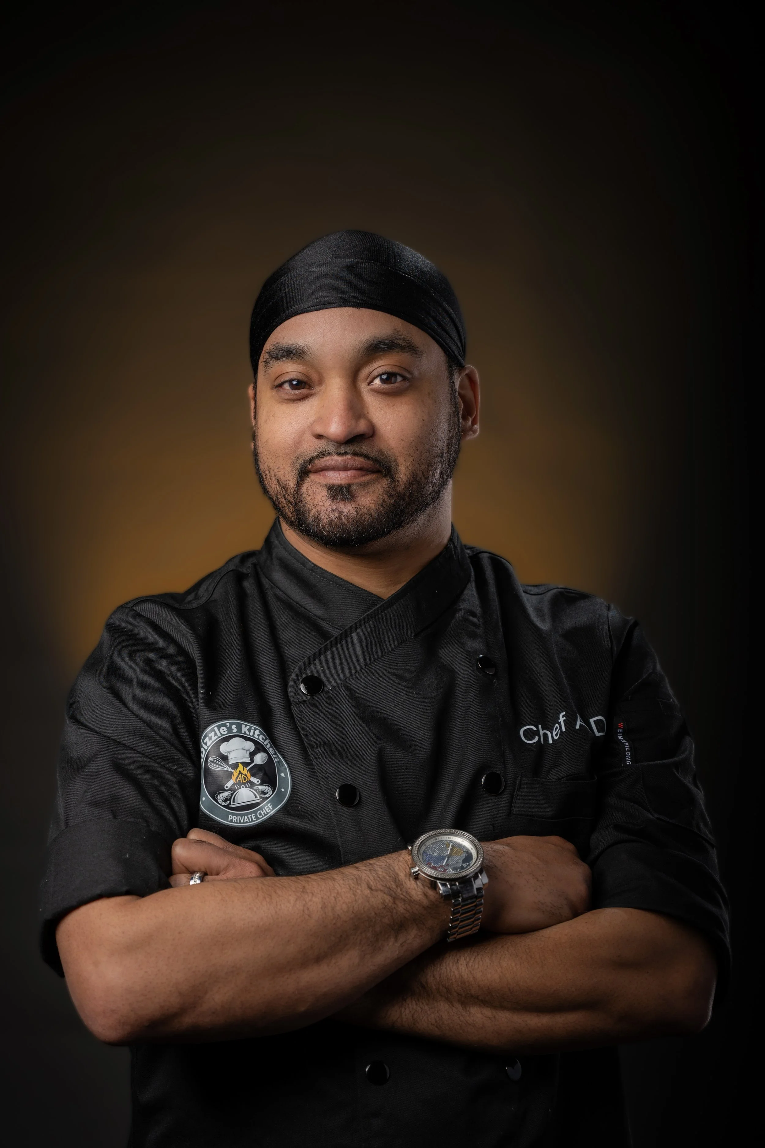 A man in a black chef's jacket with a patch that reads 'Dizzle's Kitchen Private Chef' and a badge on the right that says 'Chef AD'. He is wearing a black head covering and a silver wristwatch, standing with his arms crossed against a dark background.