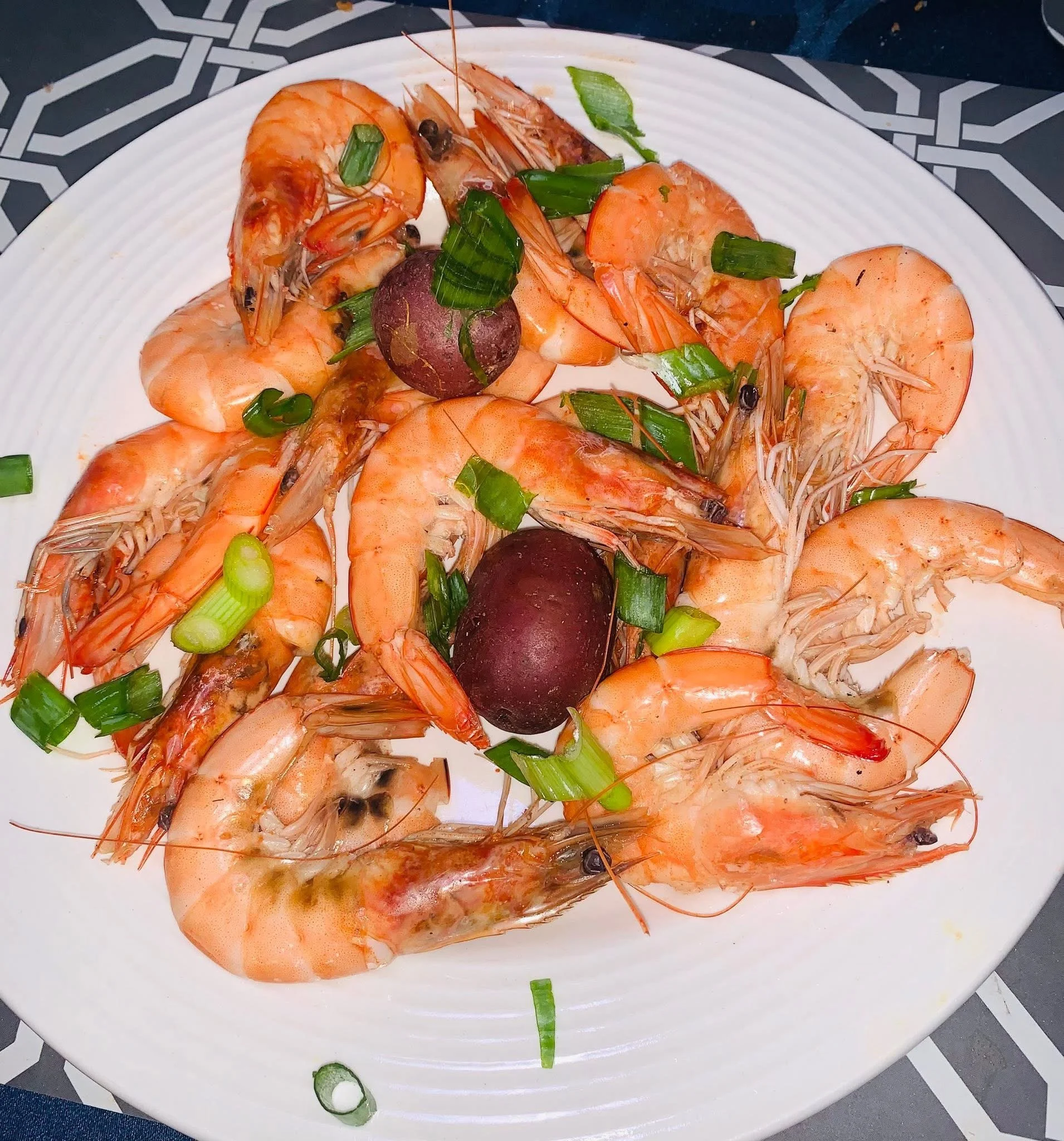 Plate of cooked shrimp garnished with chopped green onions and slices of radish, on a decorative black and white tablecloth.