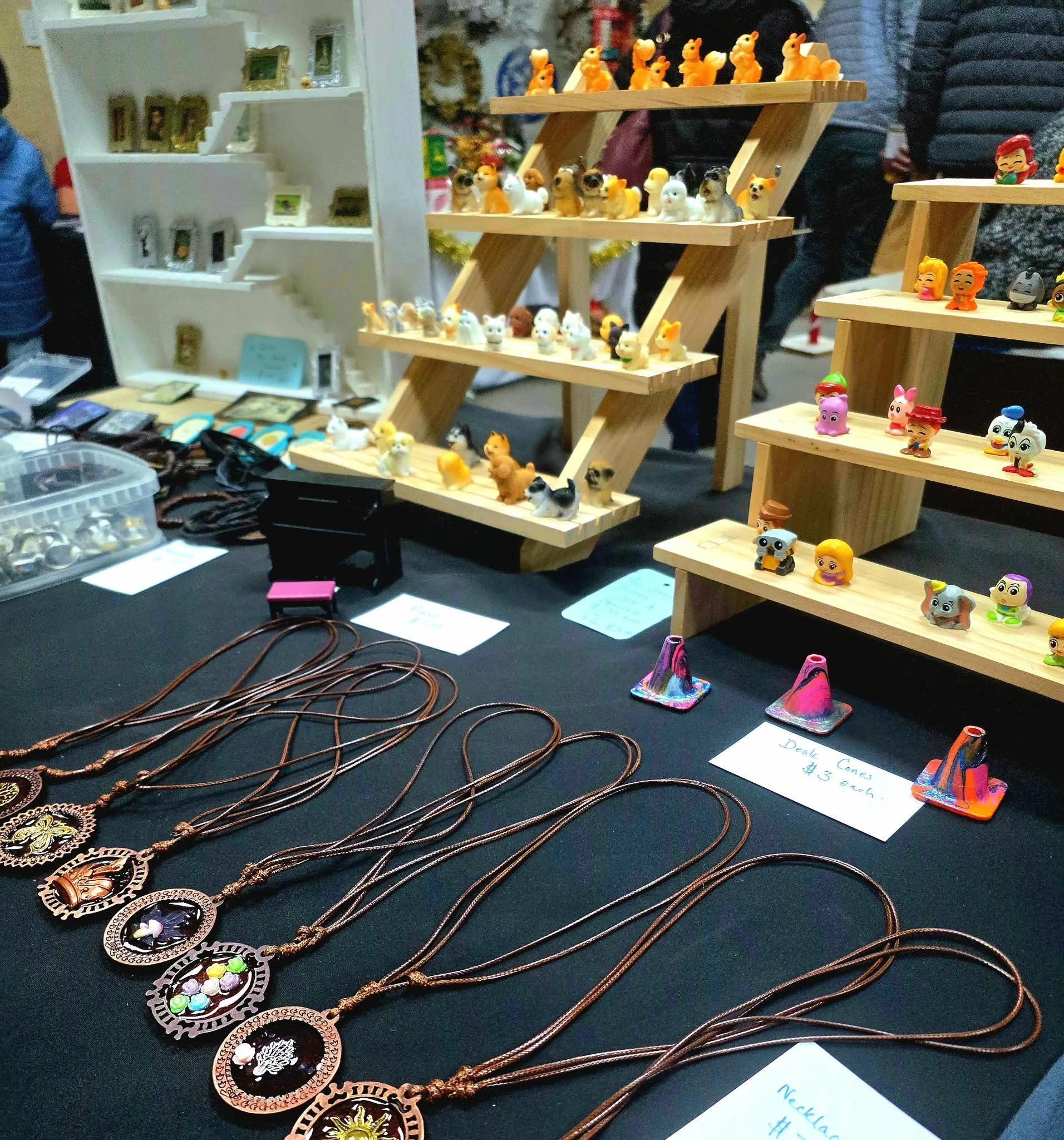 Display of handmade jewelry including necklaces with circular pendants featuring intricate designs, arranged on a black table at a market or craft fair. Several shelves showcase various small figurines and decorative items, and people are visible in the background.