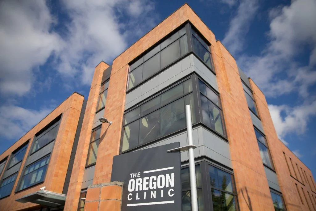 View of a modern multi-story building with brick and glass exterior, sign reading "The Oregon Clinic," cloudy sky background.