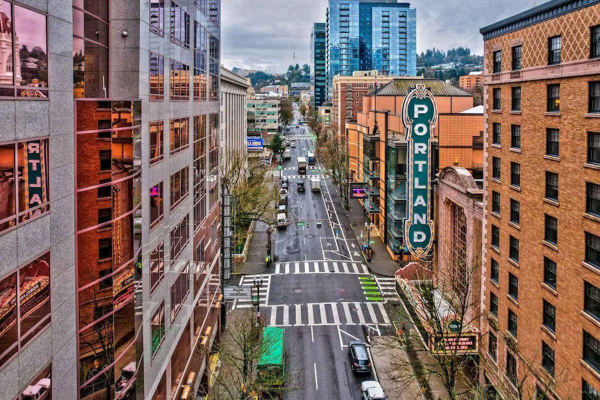 View of downtown Portland, Oregon from above, showing city streets with parked cars, crosswalks, and buildings, including a prominent Portland sign, on a cloudy day.