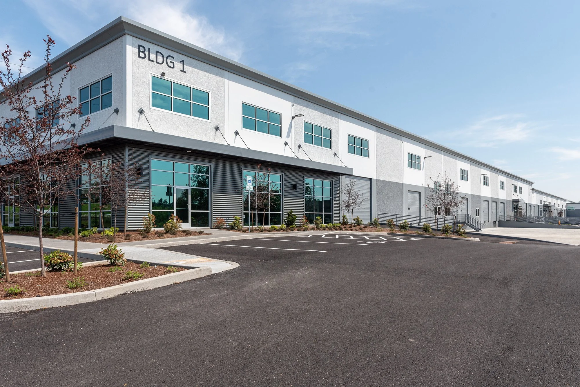Exterior of a modern industrial warehouse building labeled 'BLDG 1', with large glass windows, landscaped trees and shrubs, and empty parking lot.