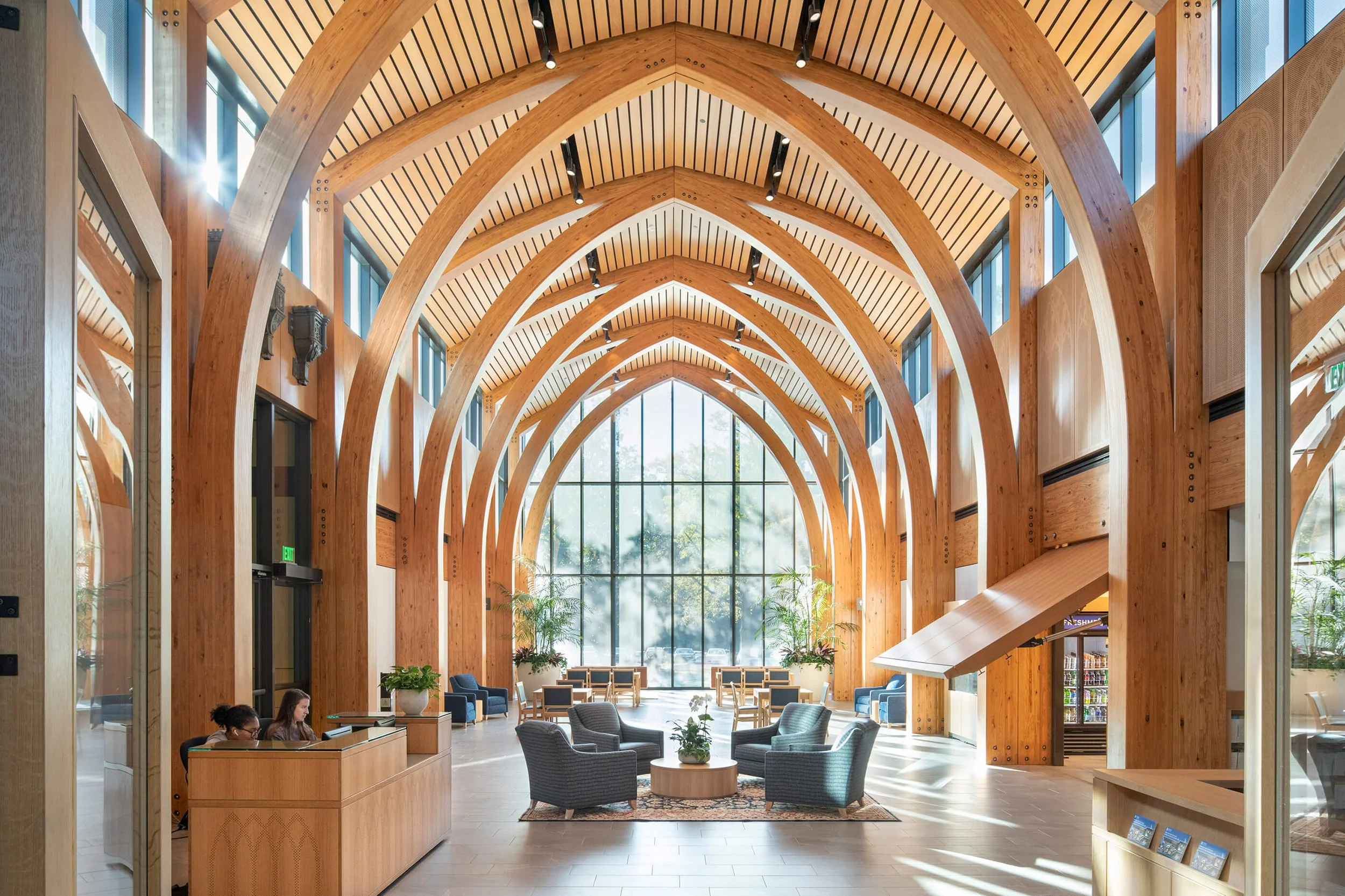 A spacious, modern lobby with tall wooden arches, large windows, and a seating area with chairs and plants.
