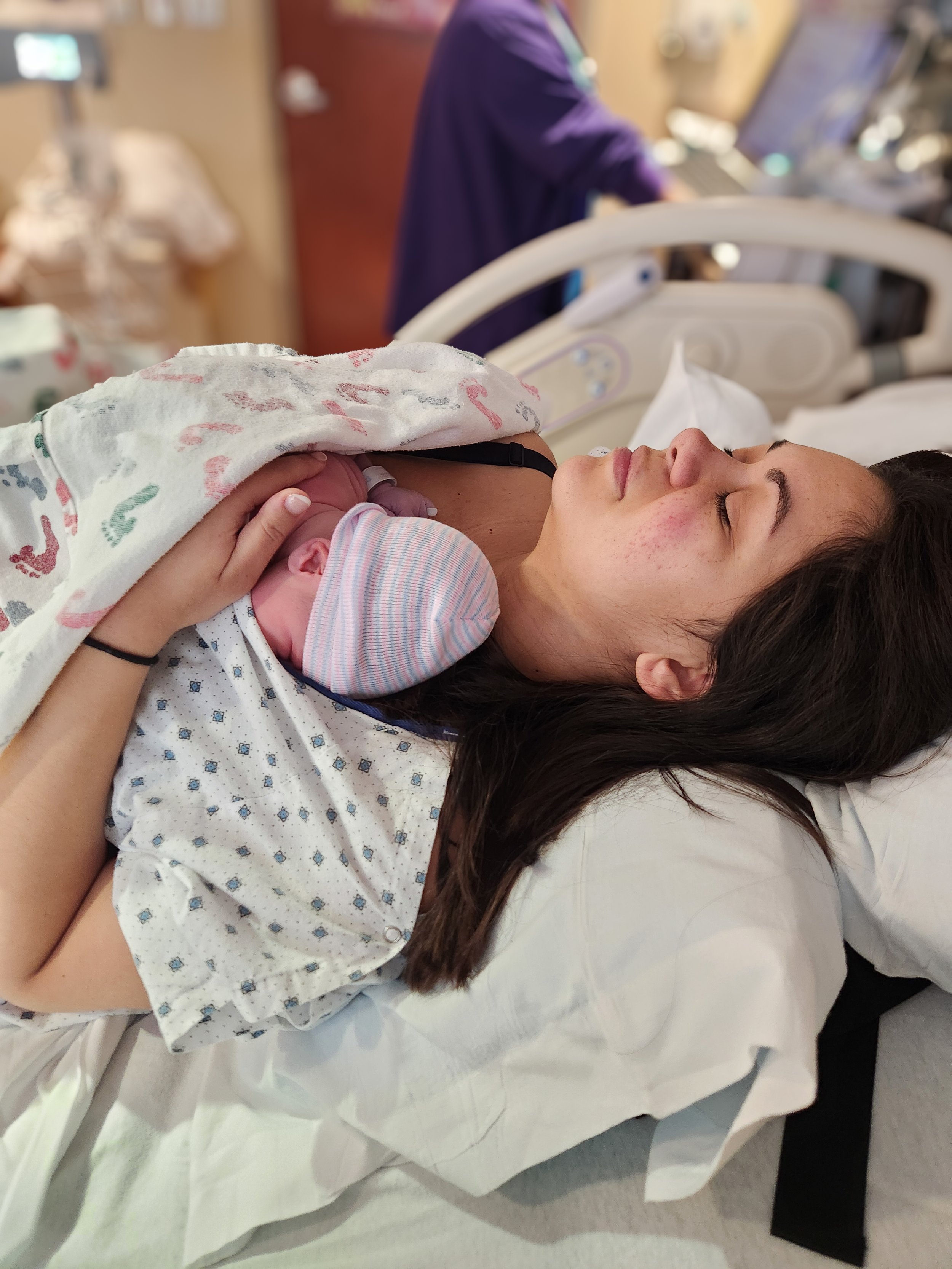 A woman lying in a hospital bed with her eyes closed, holding a newborn baby against her chest. The woman appears to be resting after childbirth.